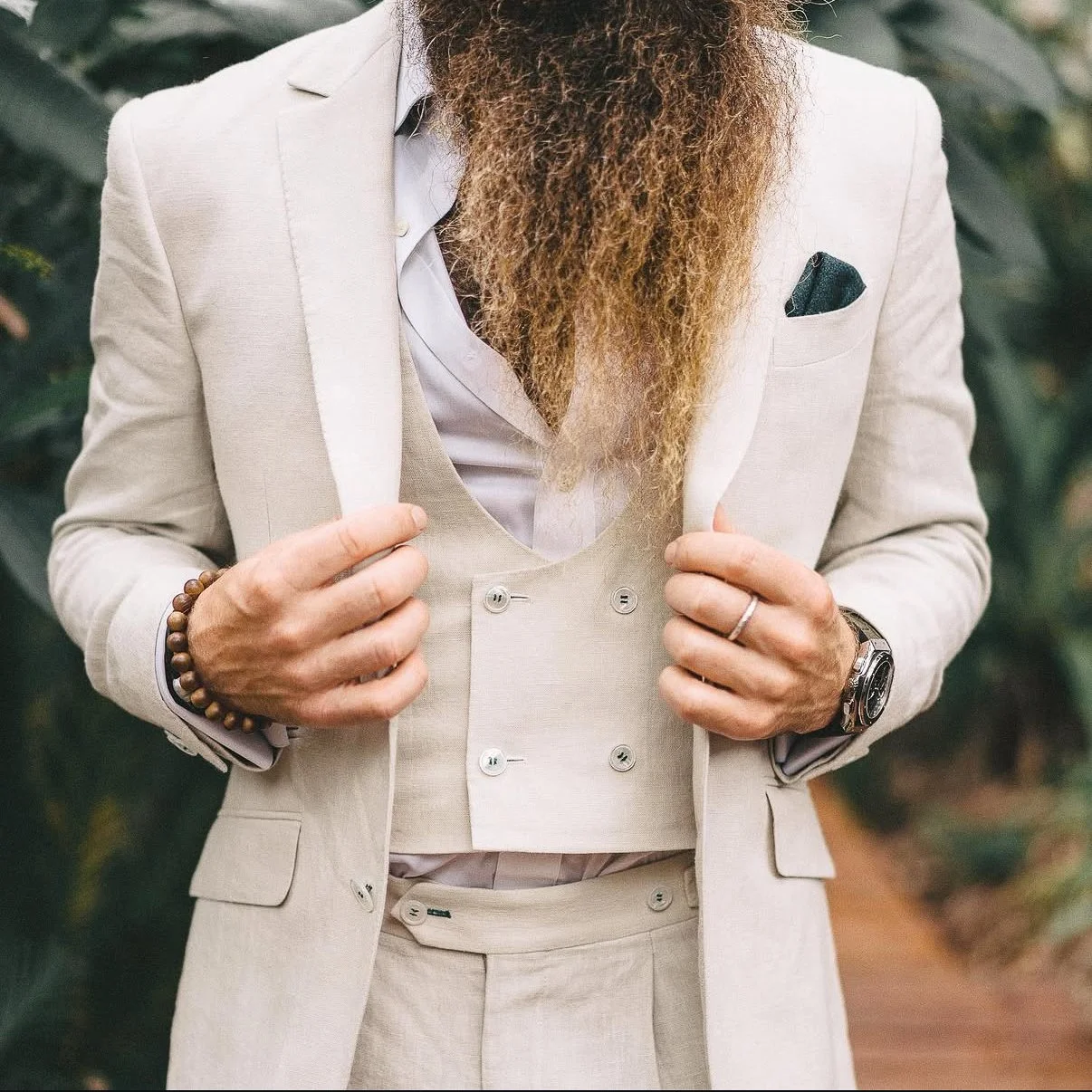 A man in a beige suit adjusting his jacket, with a long beard, wearing a white shirt, a dark pocket square, a beaded bracelet, a wristwatch, and a ring.