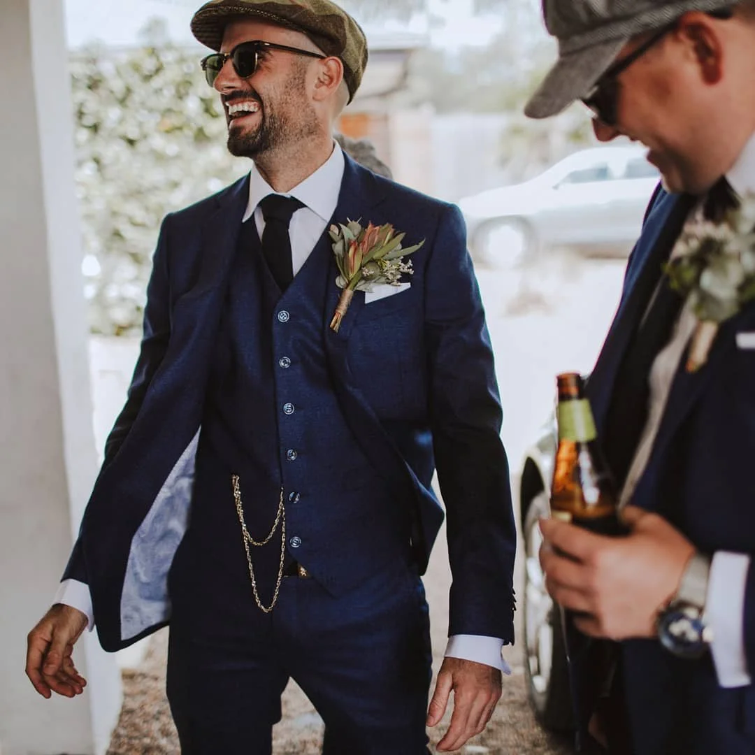 Two men dressed in dark blue suits with boutonnières, celebrating at a wedding or special occasion, outdoors with natural light.