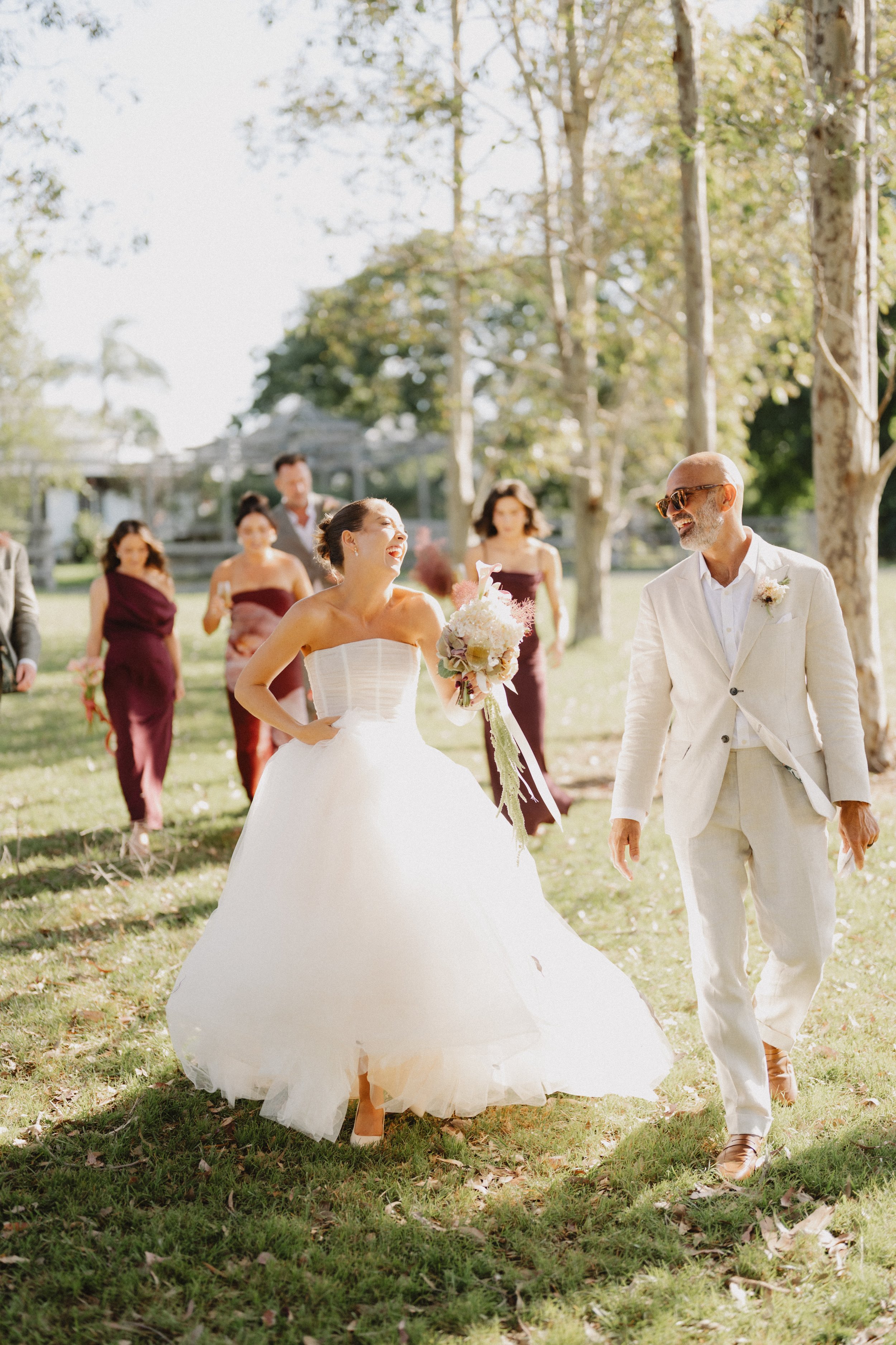 A wedding celebration outdoors with a bride in a white gown holding a bouquet, and an older man in a light suit and sunglasses, surrounded by bridesmaids in burgundy dresses, trees, and a gazebo in the background.