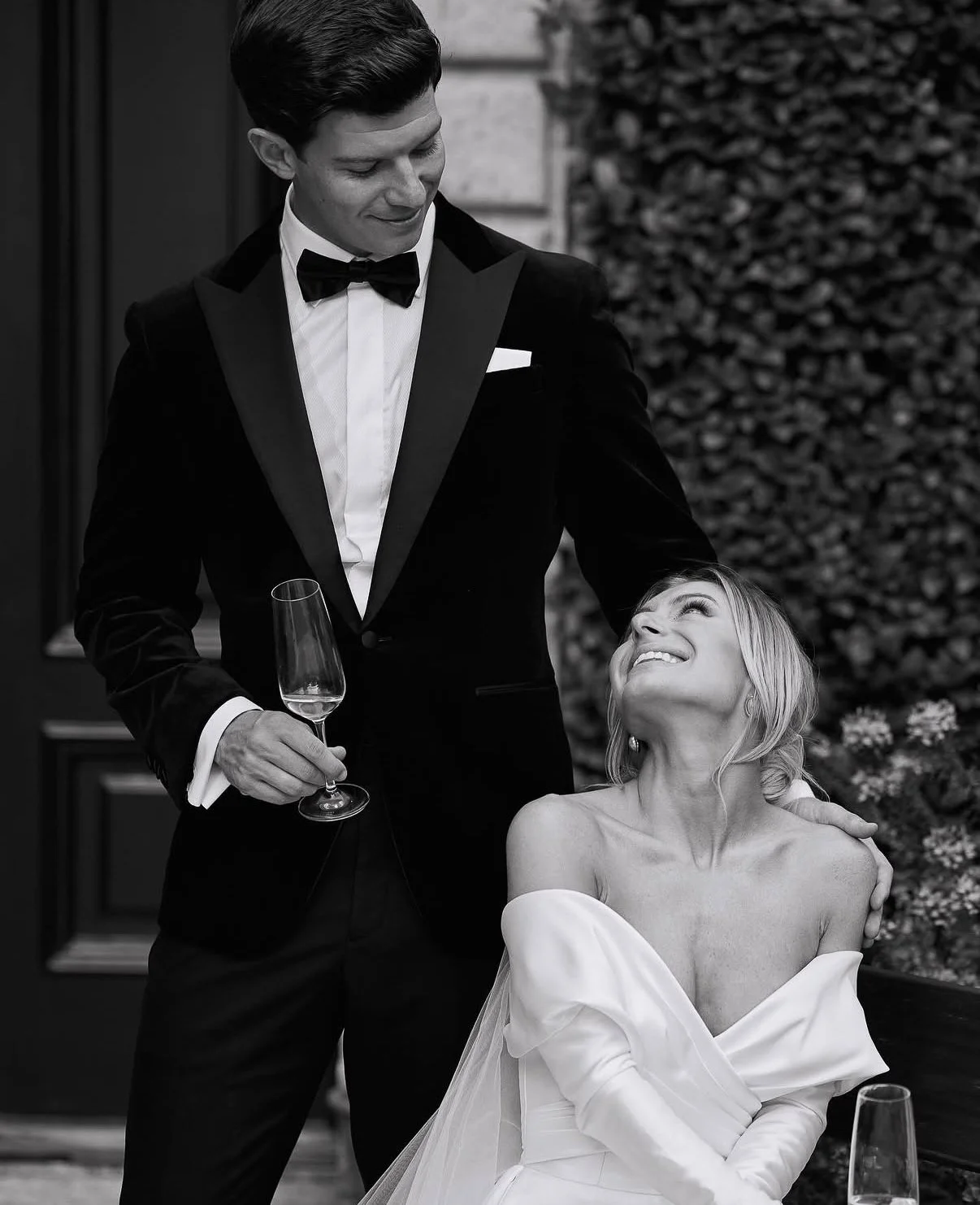 A black and white photo of a man in a tuxedo holding a glass of champagne, standing next to a woman in a strapless dress with a smile, looking up at him. They are outdoors with a textured wall and foliage in the background.