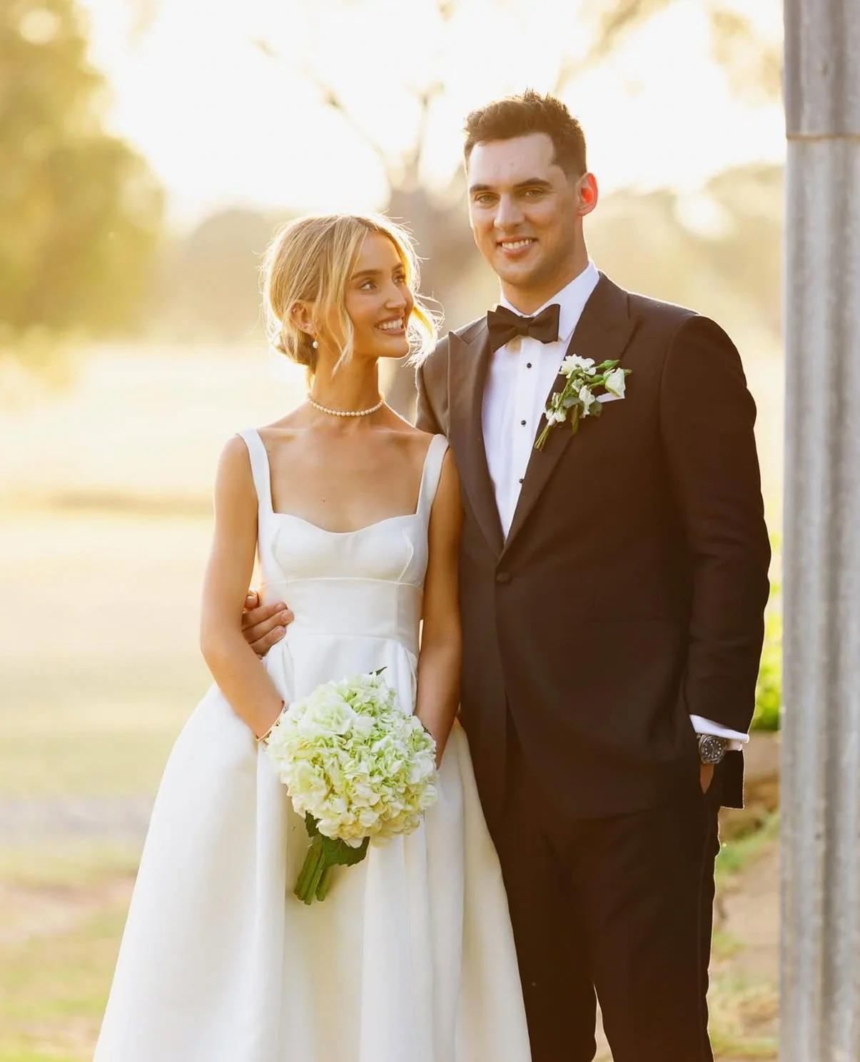 A bride and groom standing outdoors during sunset, smiling, dressed in wedding attire. The bride wears a white gown and pearl necklace, holding a bouquet of white flowers. The groom wears a black tuxedo with a bow tie and boutonniere.