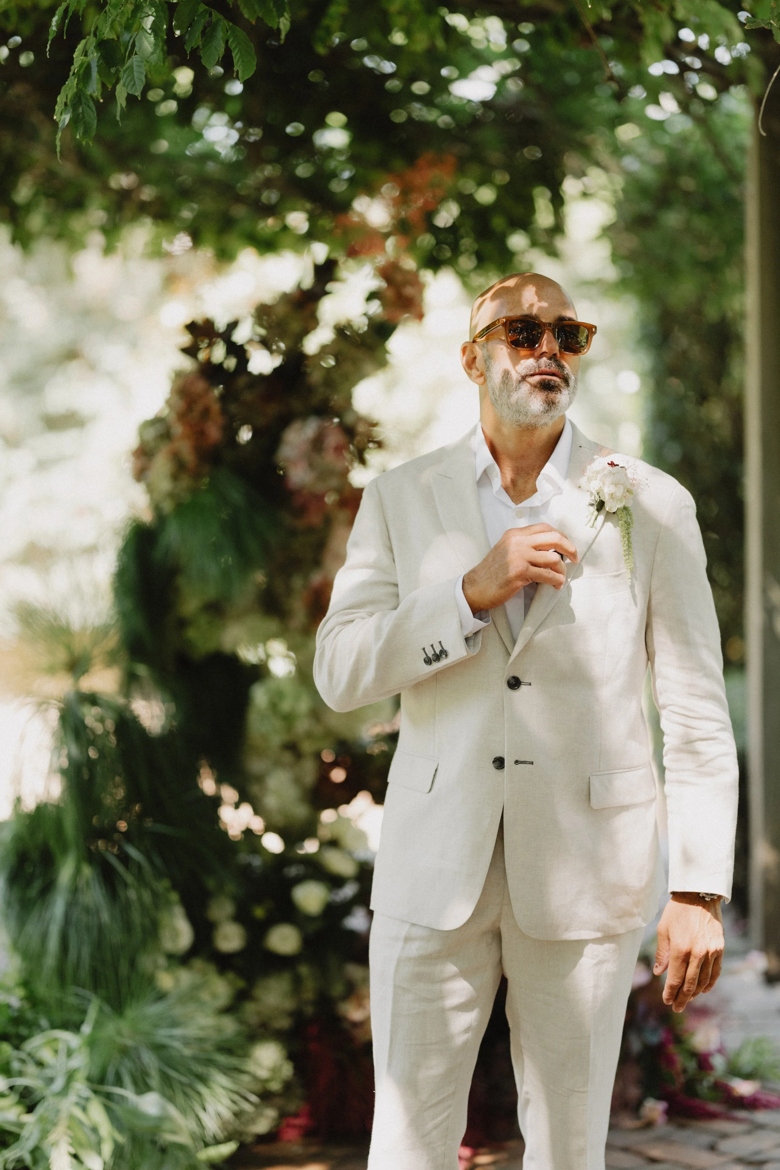 A man in a cream-colored suit with sunglasses standing outdoors among greenery and flowers.