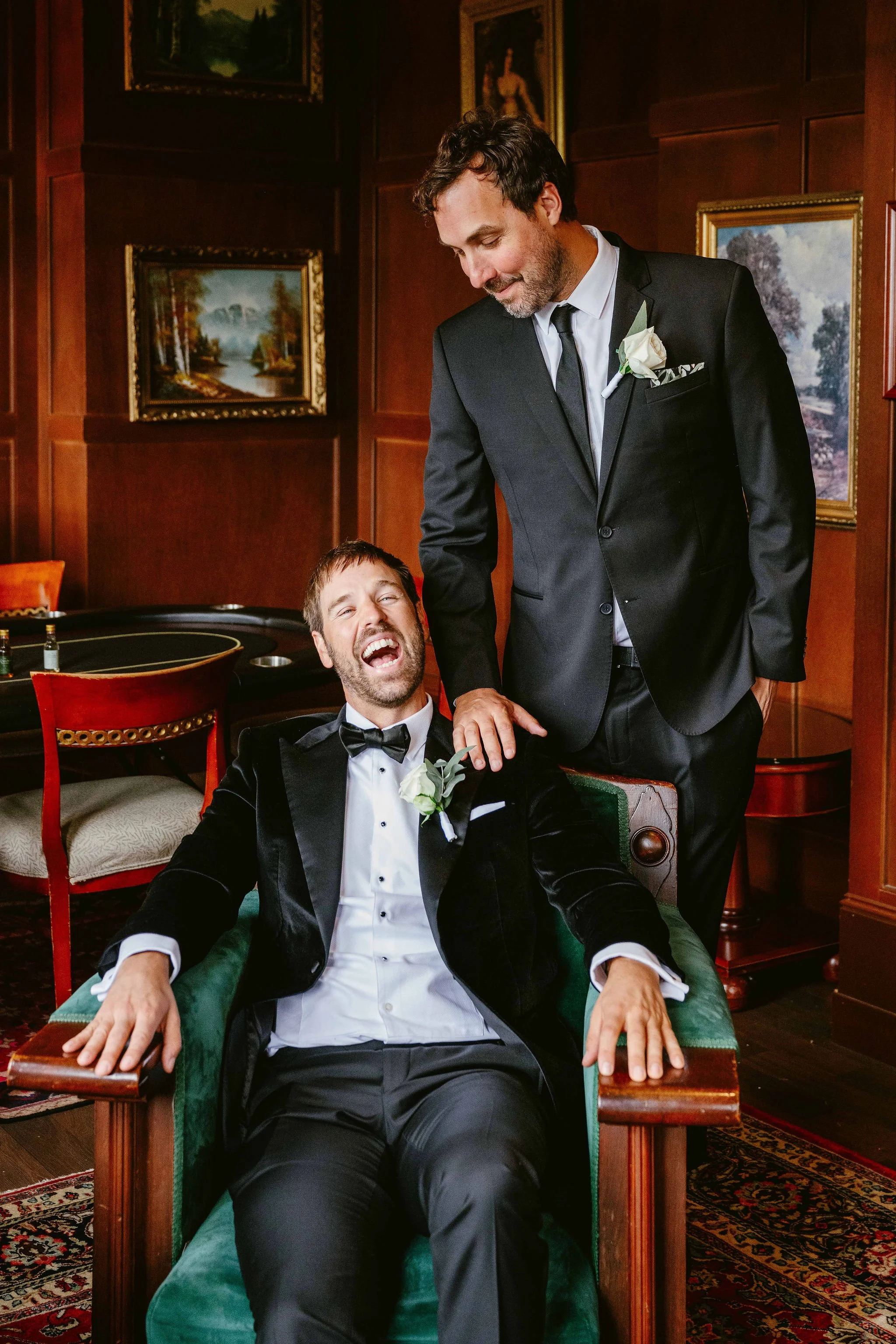 Two men dressed in tuxedos in a vintage room with framed landscape paintings. One man is seated in a green velvet chair with his hands on armrests, laughing, while the other man stands beside him, smiling and holding his hand.