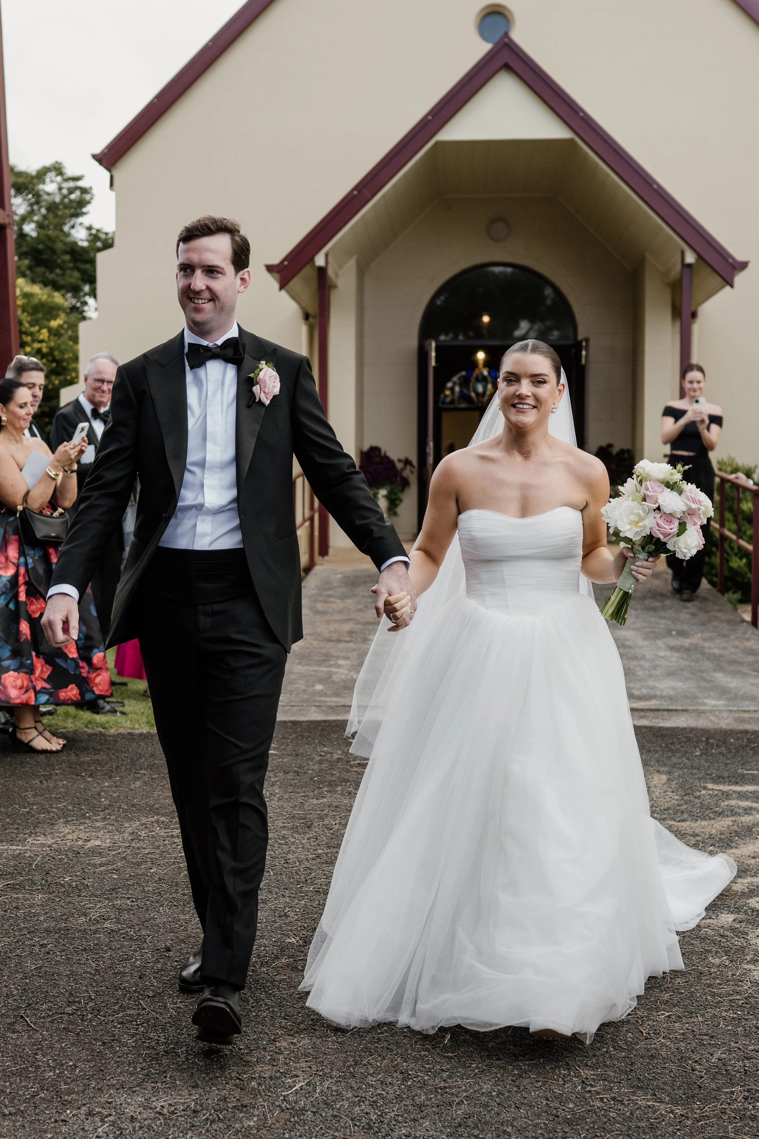 The bride and groom, both smiling, walk hand in hand outside a church, surrounded by guests taking photos.