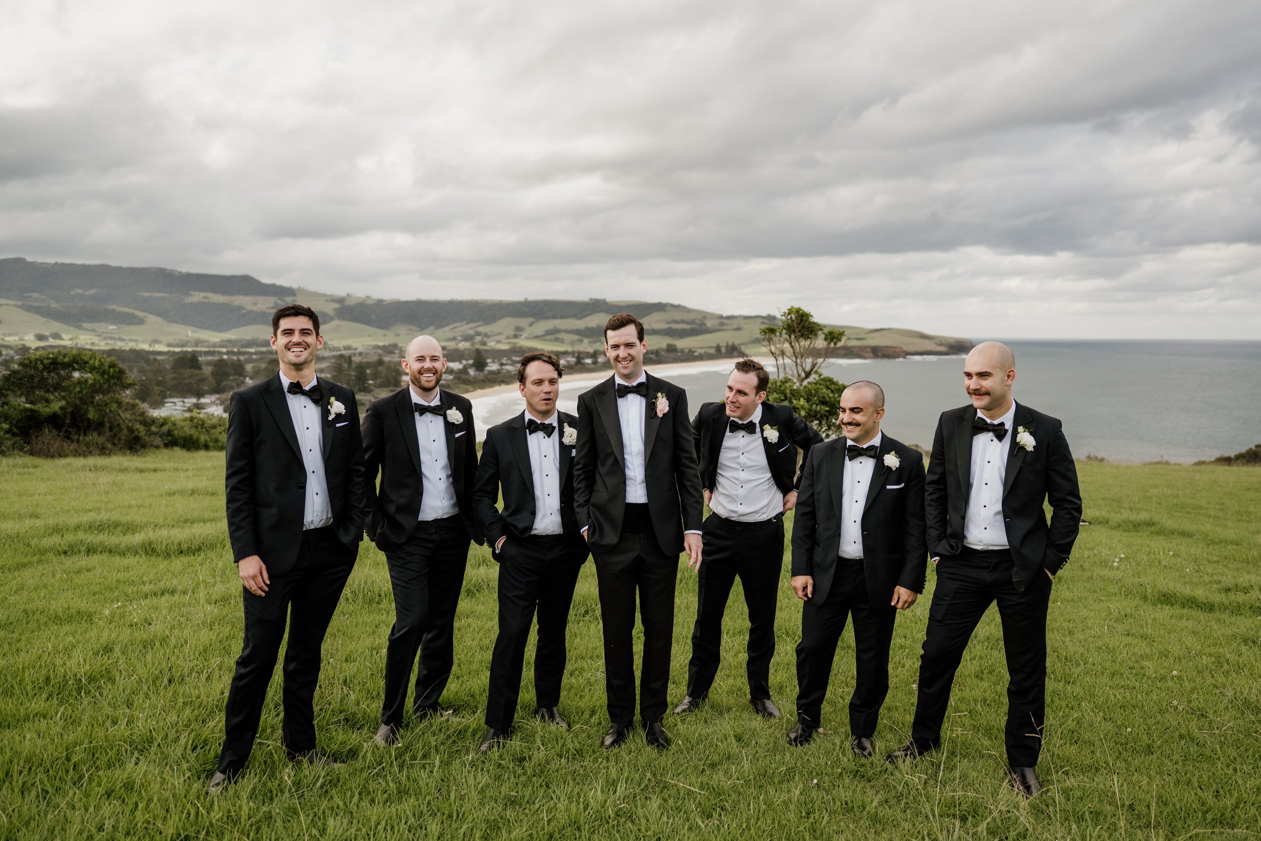 A group of seven men in tuxedos and bow ties standing on a grassy hilltop near the coast, with a cloudy sky overhead and rolling green hills in the background.