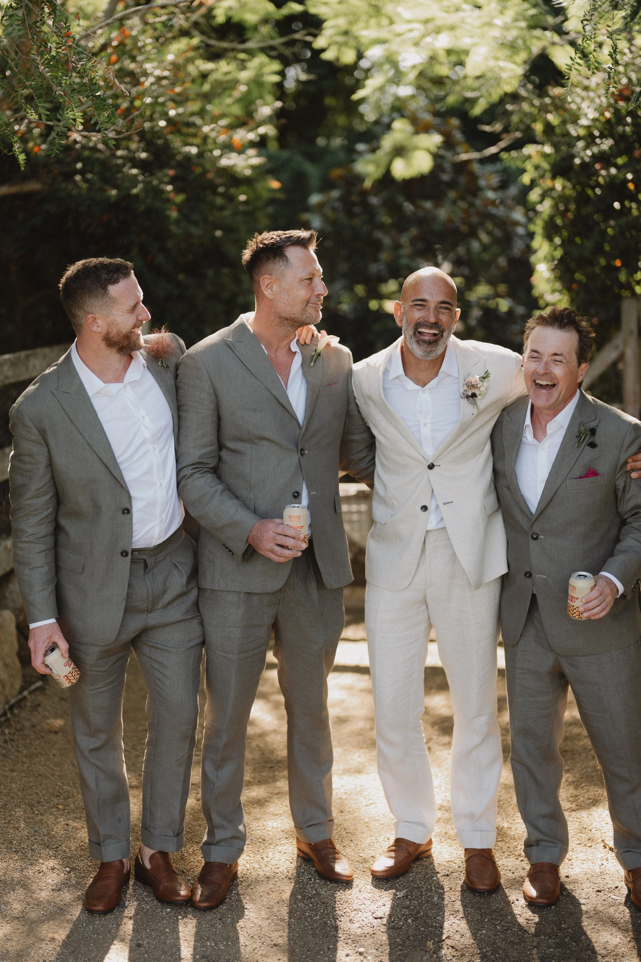 Four men dressed in suits, glasses of drinks in hand, standing together outdoors, smiling and laughing in a park or garden setting with greenery and sunlight.