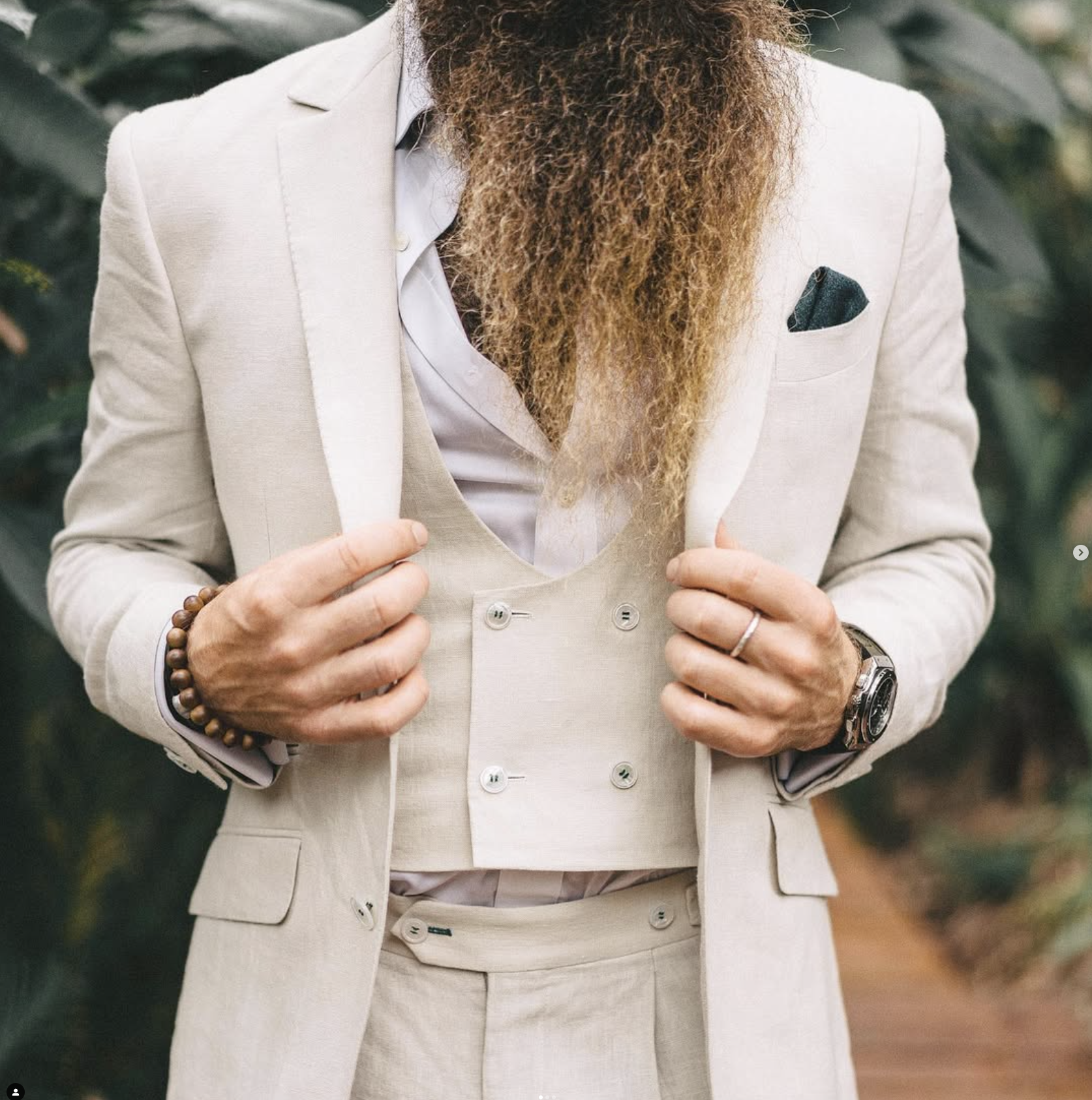 A person dressed in a cream-colored suit with a white shirt, holding the lapel of their blazer. The person has a full, curly beard and is wearing a wristwatch and a beaded bracelet. The background is blurred with green foliage.