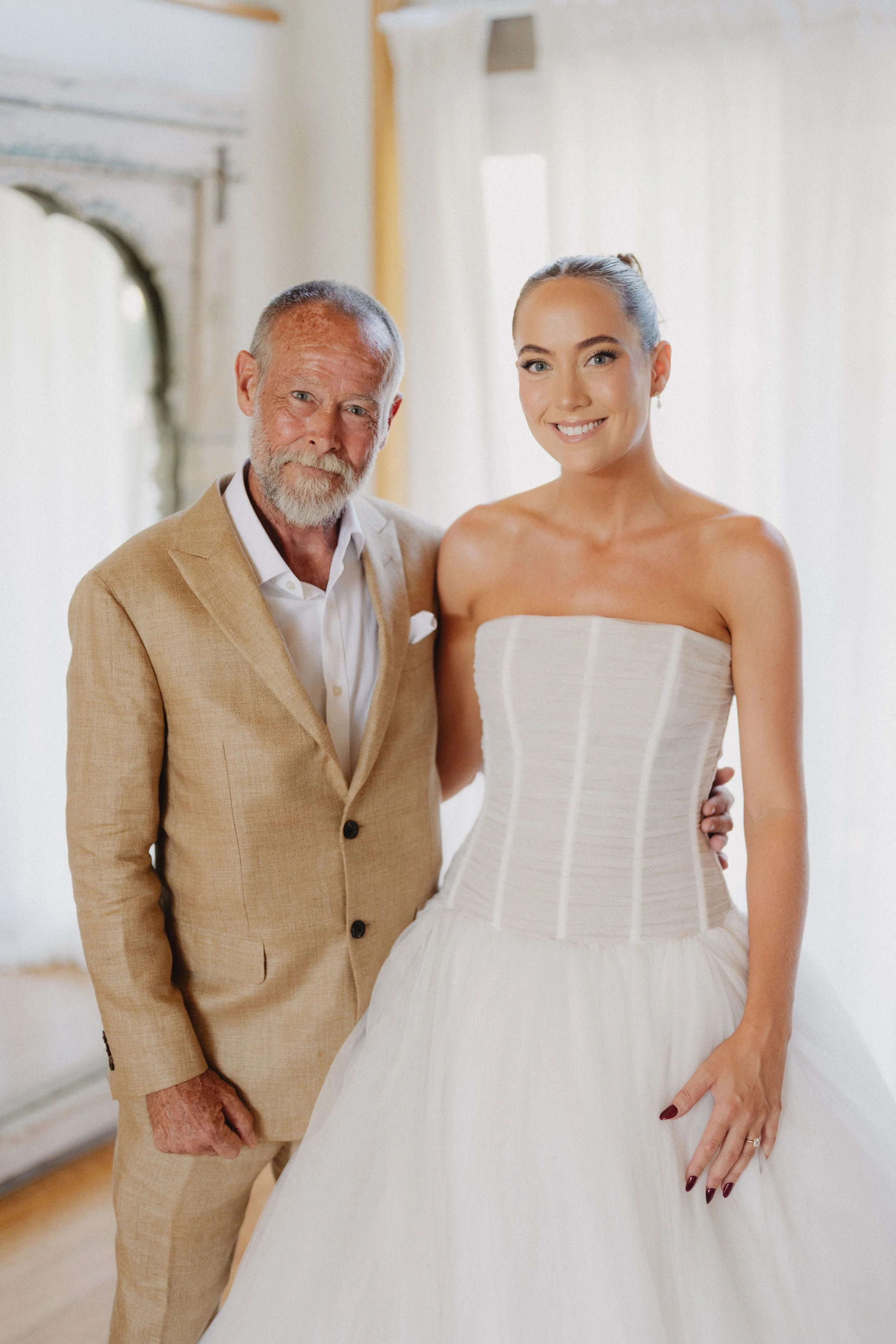 An elderly man and a young woman in a wedding dress pictured together indoors, smiling at the camera.