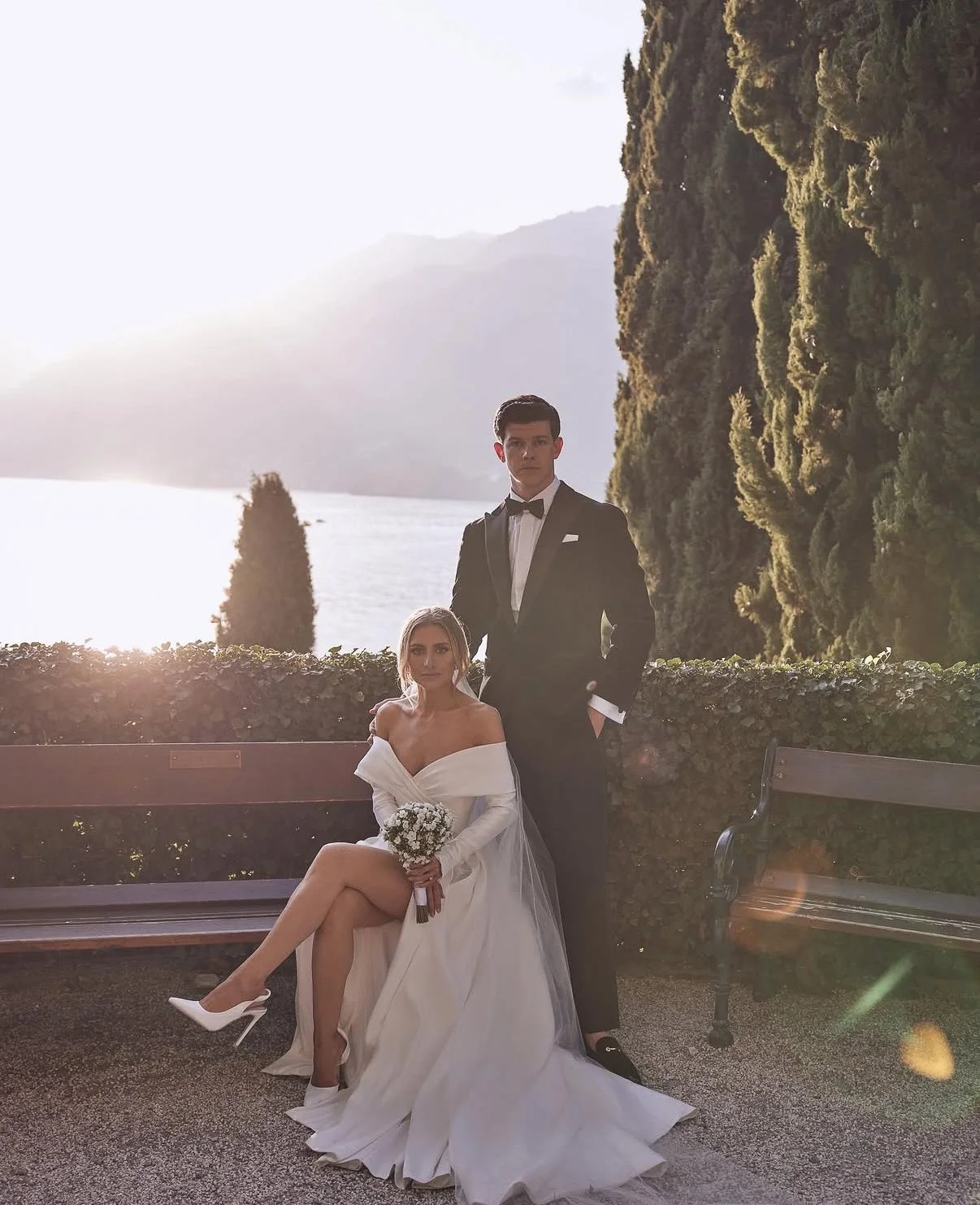 A bride and groom dressed in wedding attire standing outdoors near a body of water with mountains in the background, during sunset.