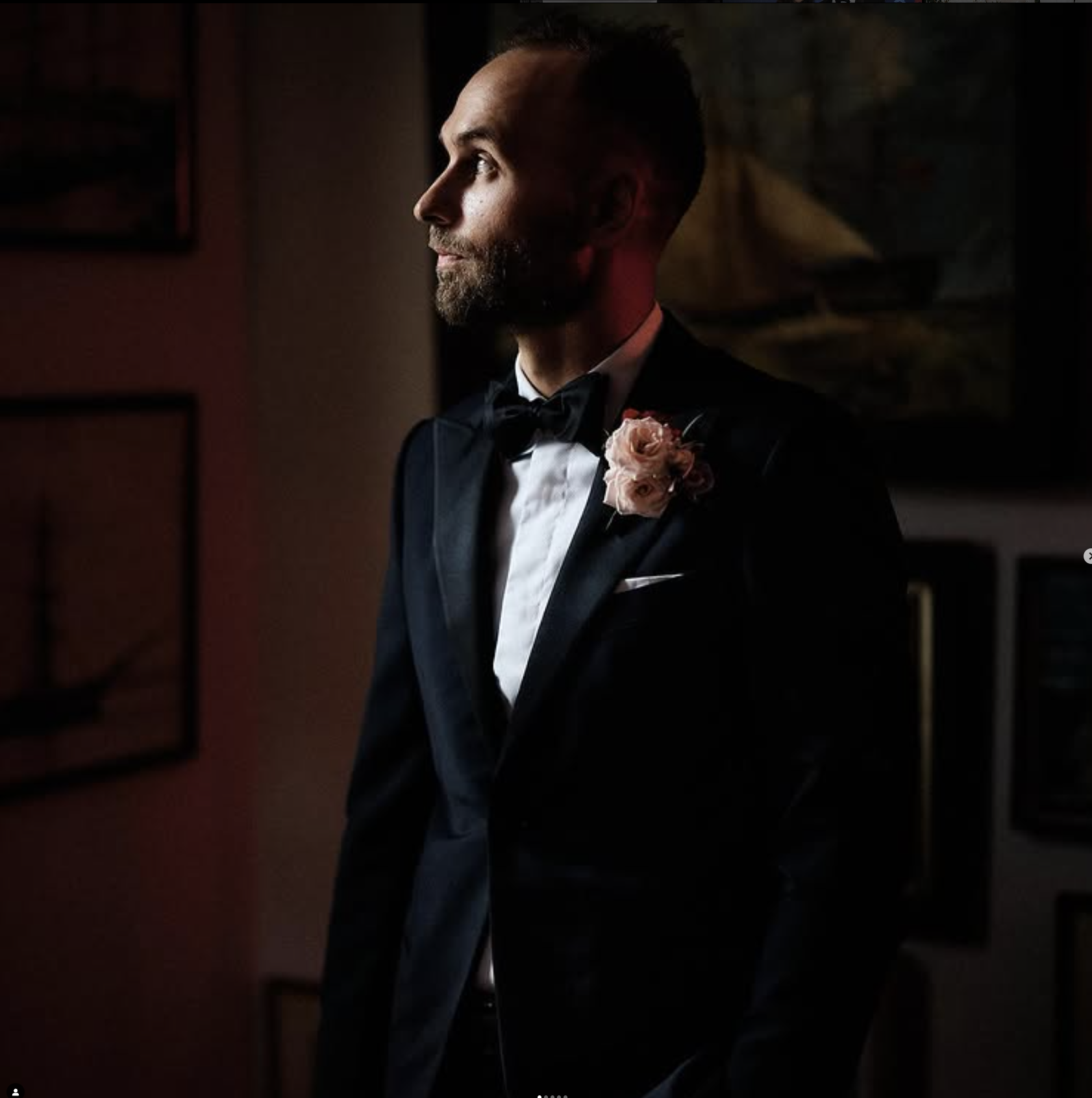 Side profile of a man dressed in a black tuxedo with a bowtie, a white shirt, and a pink flower boutonniere, standing indoors in dim lighting.