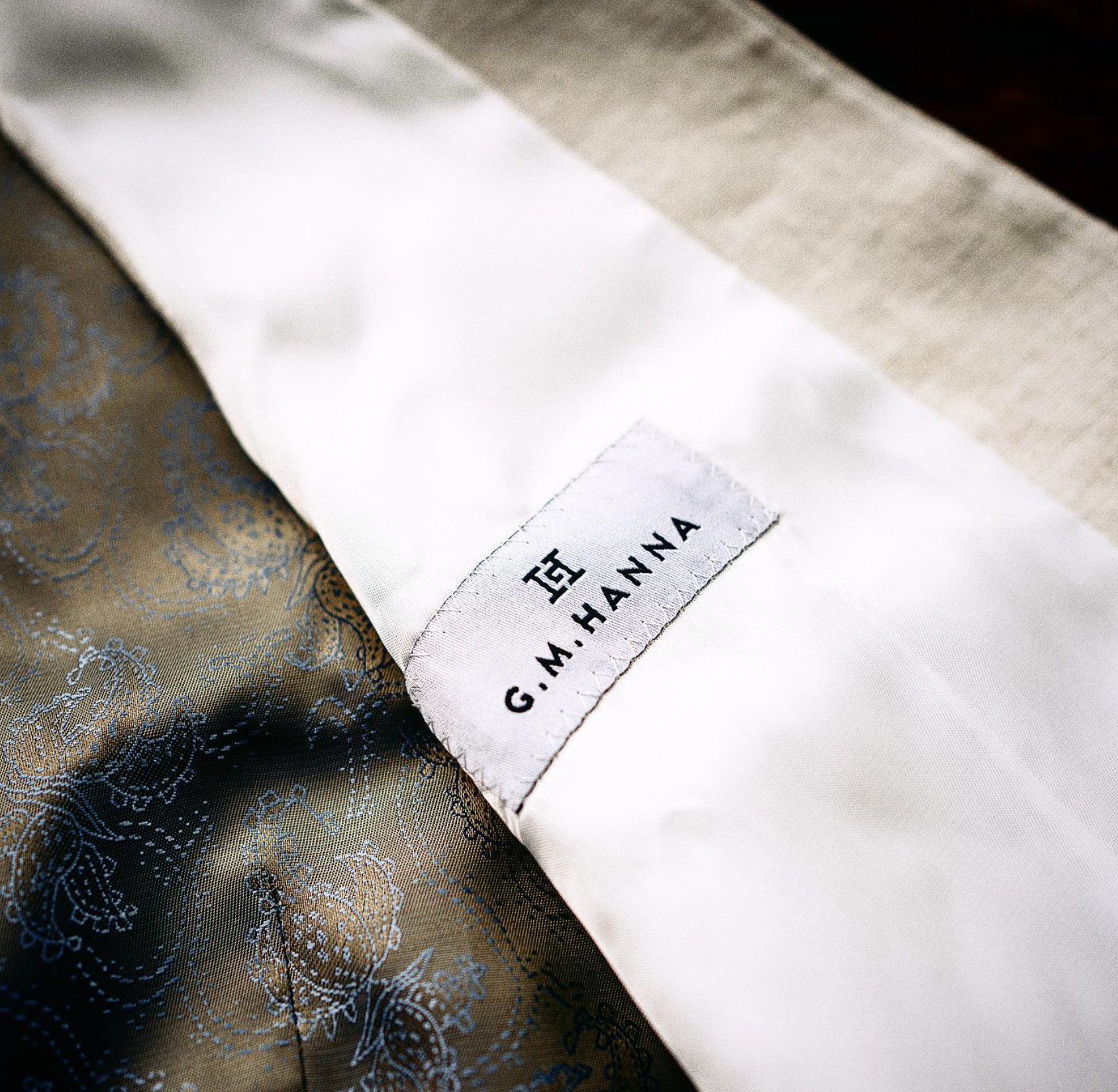 Close-up of a fabric label on a garment with the words 'G.M. HAINA' visible, attached to a piece of white fabric with a luxurious, brocade fabric underneath.