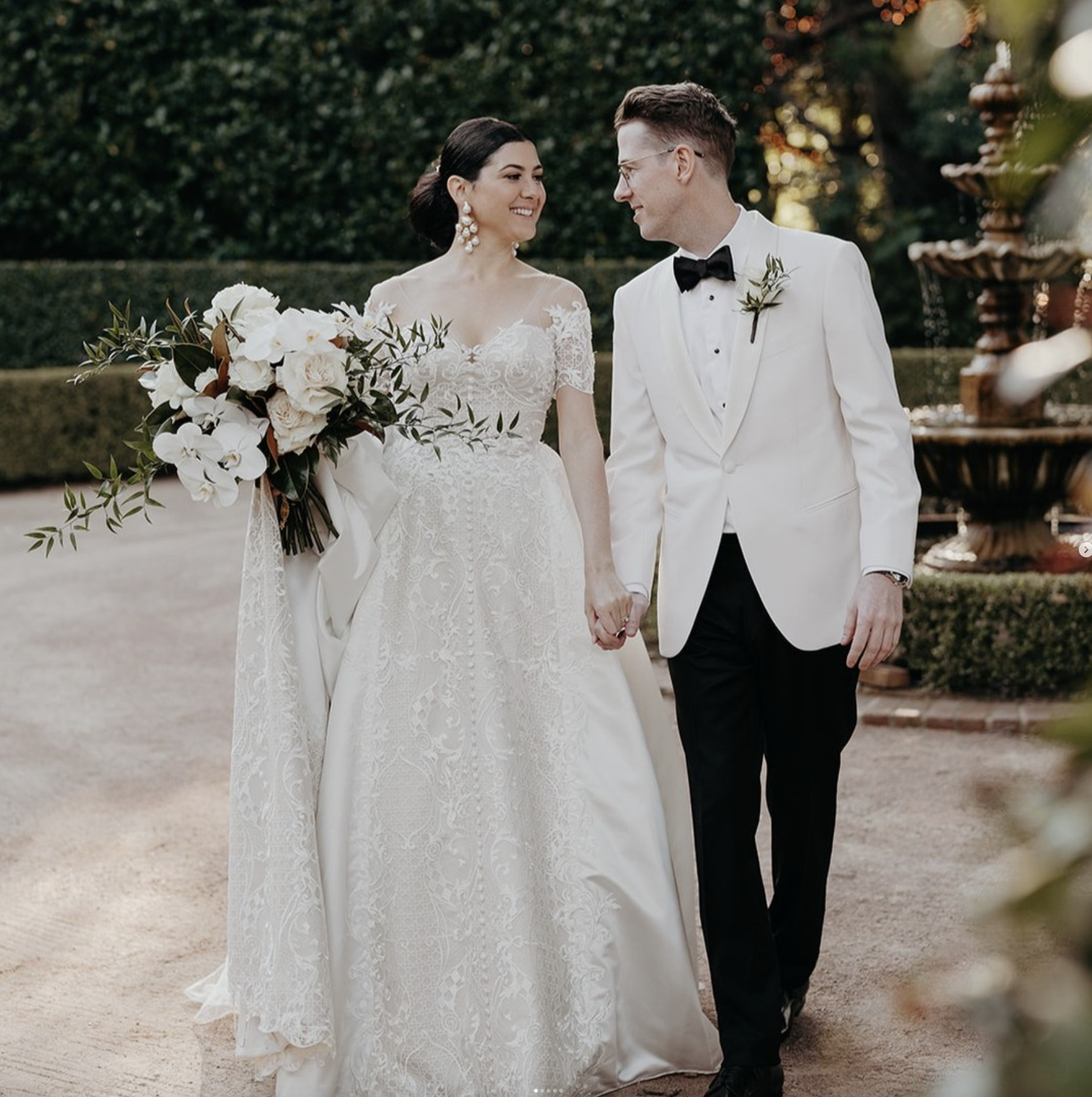 A bride and groom standing outdoors in a garden, holding hands, smiling at each other during their wedding. The bride is wearing a lace wedding dress with an off-the-shoulder neckline, large pearl earrings, and is holding a bouquet of white flowers a
