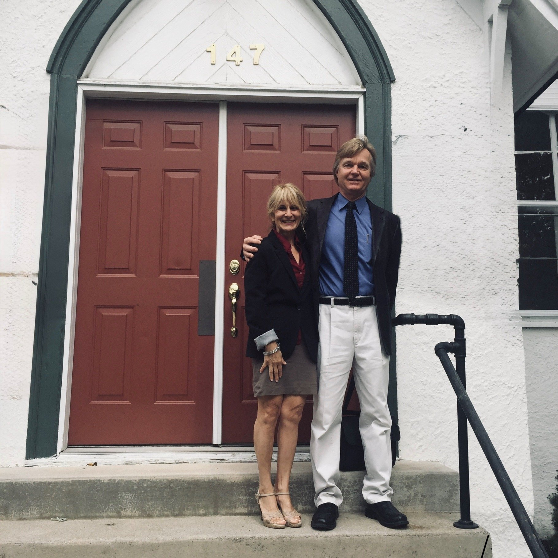 A smiling man and woman stand together on a front porch in front of a red door with the number 147 above it. The woman is wearing a black blazer, red blouse, gray skirt, and beige high heels. The man is dressed in a black blazer, blue shirt, and white pants. They are posing for the photo with their arms around each other.
