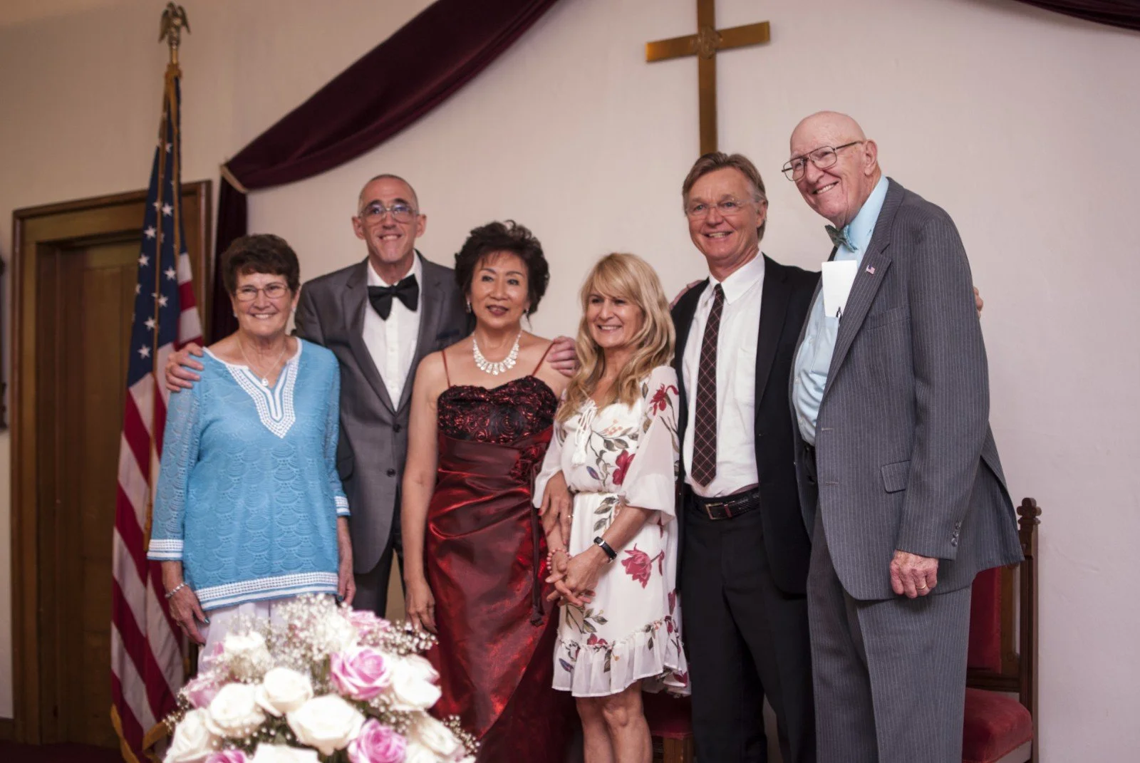 Six people standing in a group, smiling, inside a room with a wooden door, American flag, and a cross on the wall. They are dressed in formal and semi-formal attire.