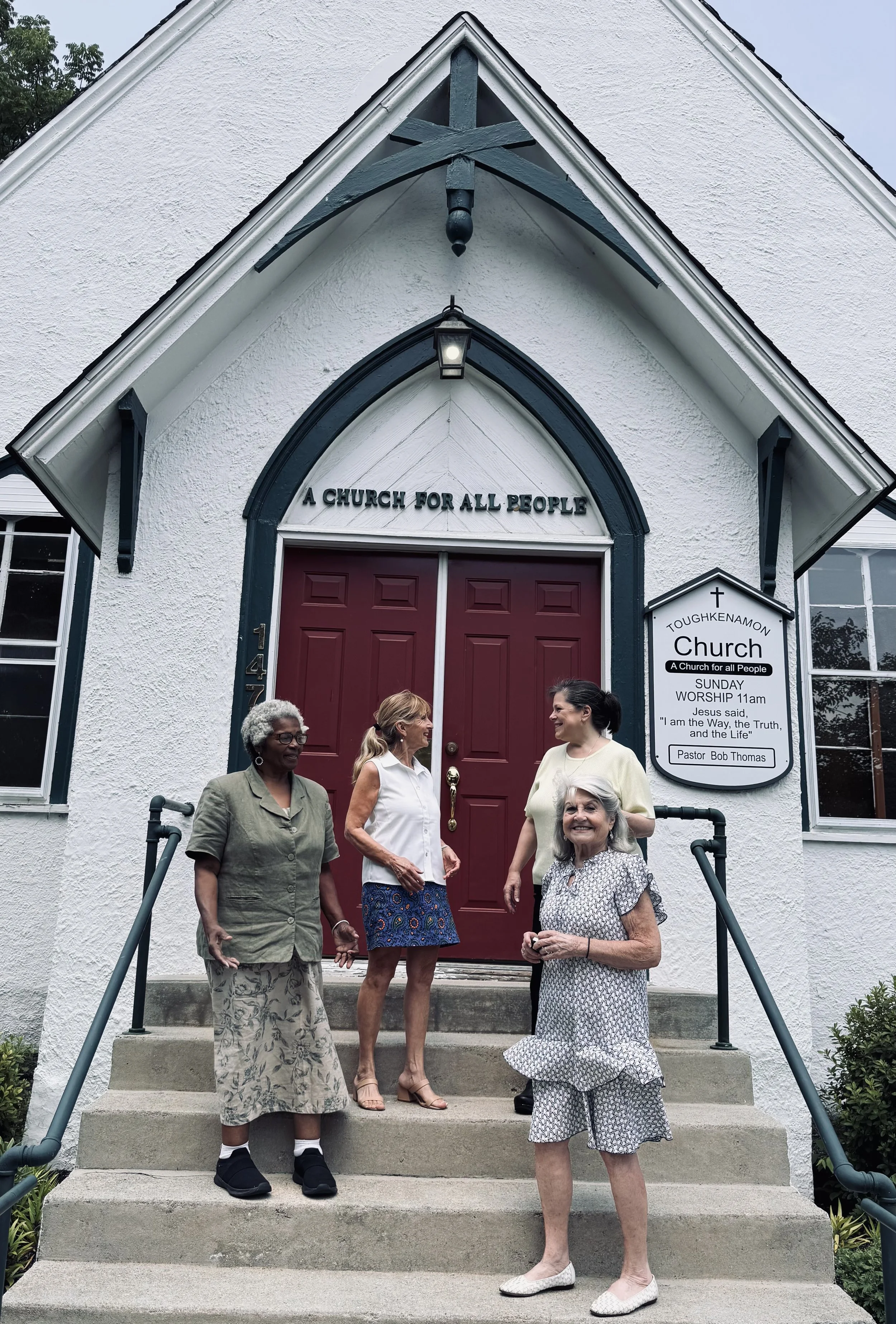 A group of five women standing on the steps of a white church with red doors, smiling and talking with each other.