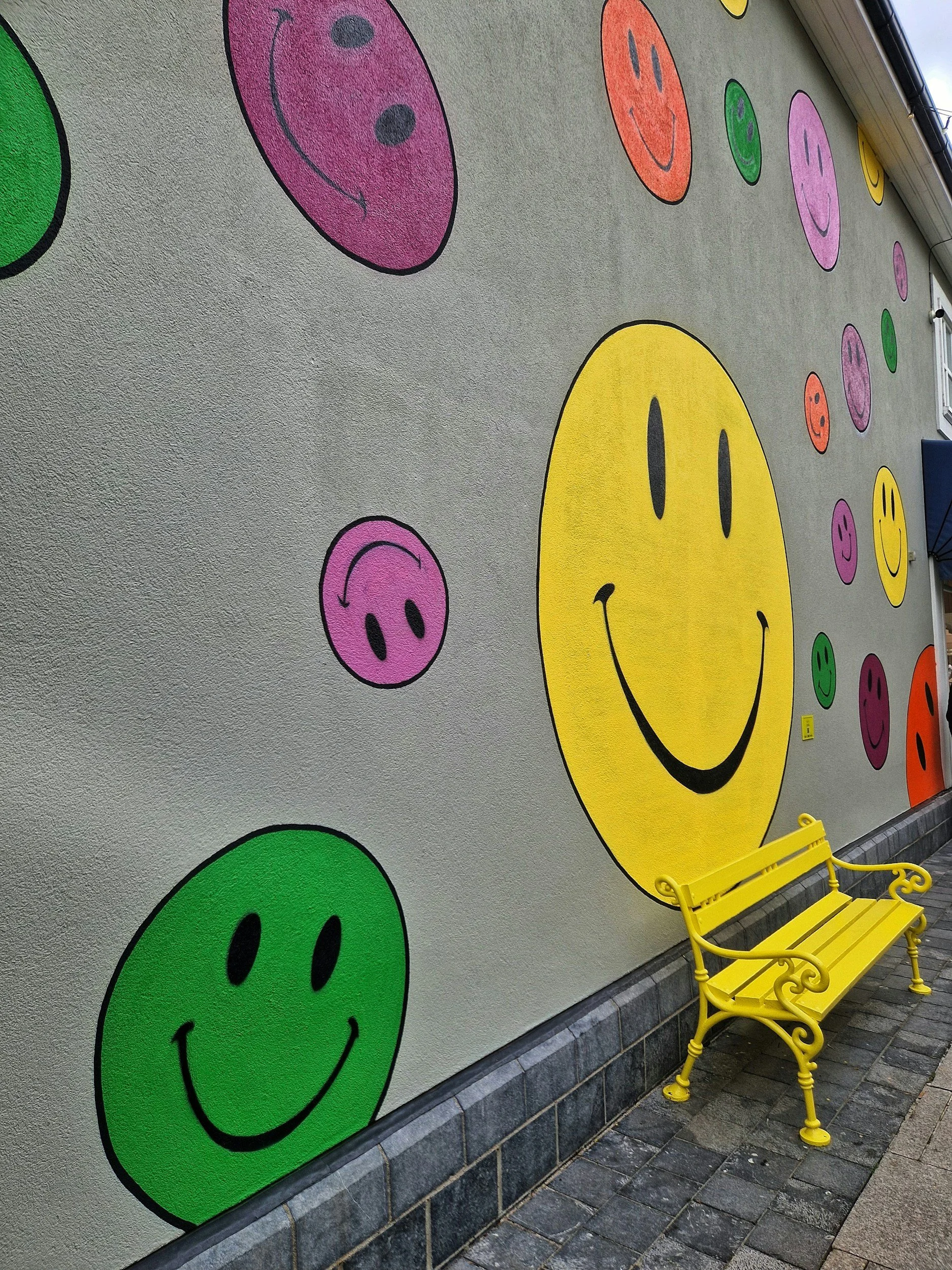 A bright yellow bench against gray wall painted with multicolored smiley faces