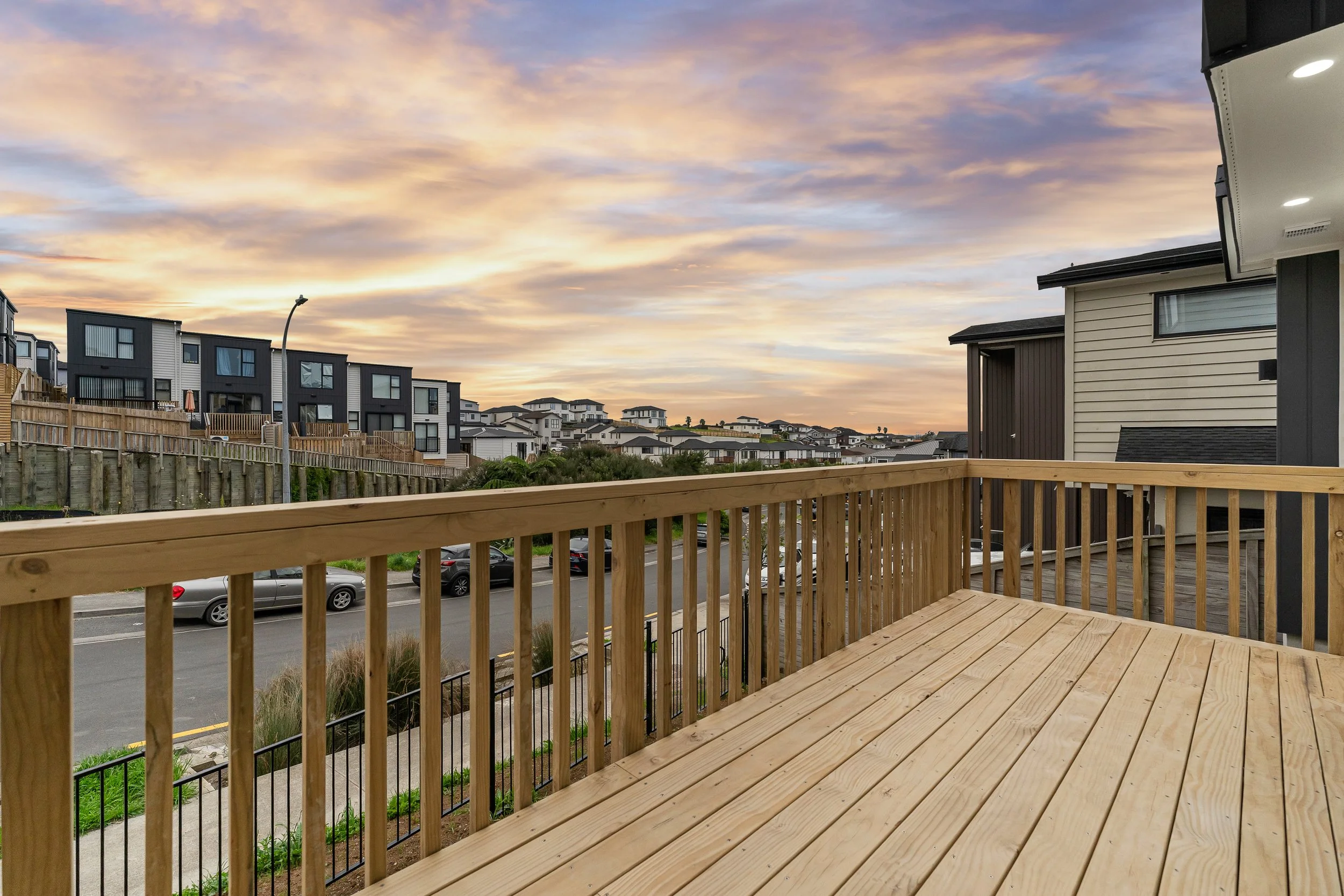 View of a suburban neighborhood with modern houses on a hill, under a colorful sunset sky, as seen from a wooden deck with railing in the foreground.
