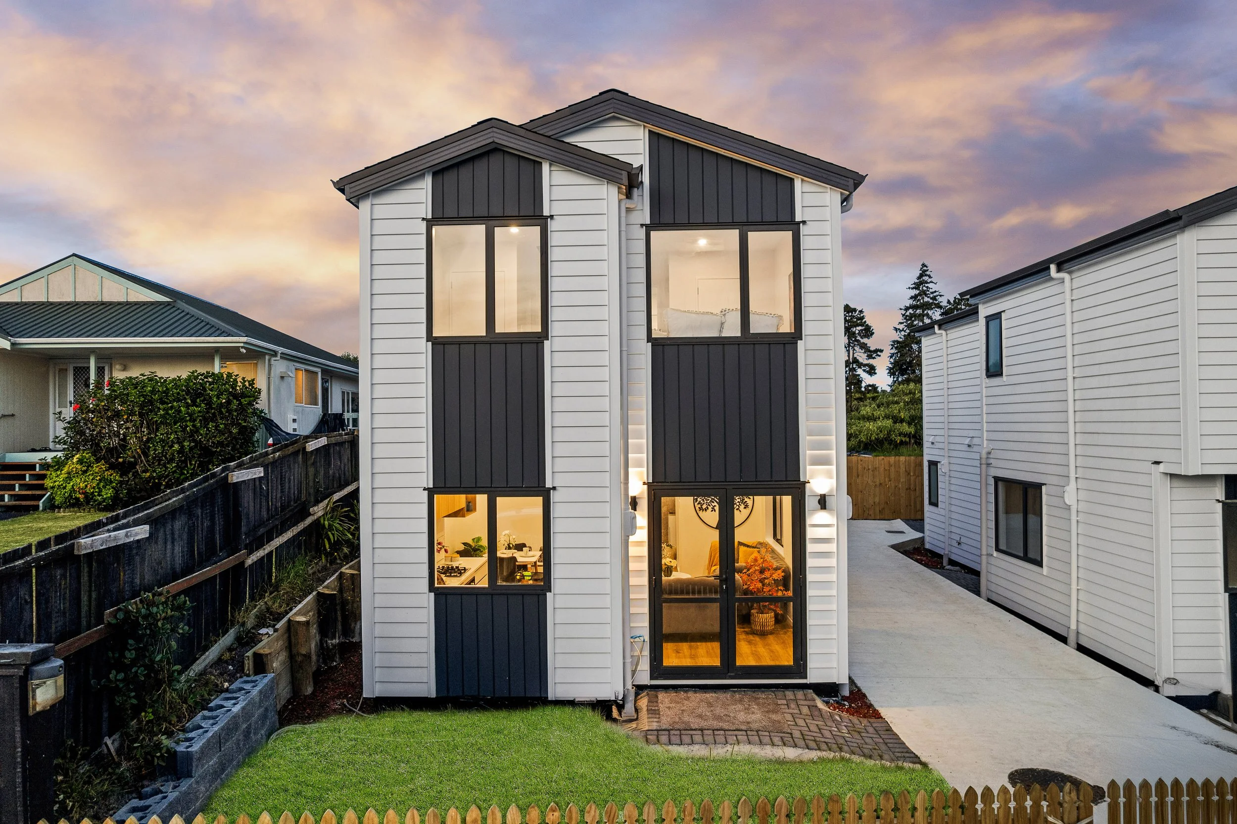 A modern house with a narrow front-view, featuring white and dark gray vertical siding, large windows, and interior lighting visible through the windows at dusk. The house is surrounded by a lawn, a wooden fence, and neighboring houses with tiled roo