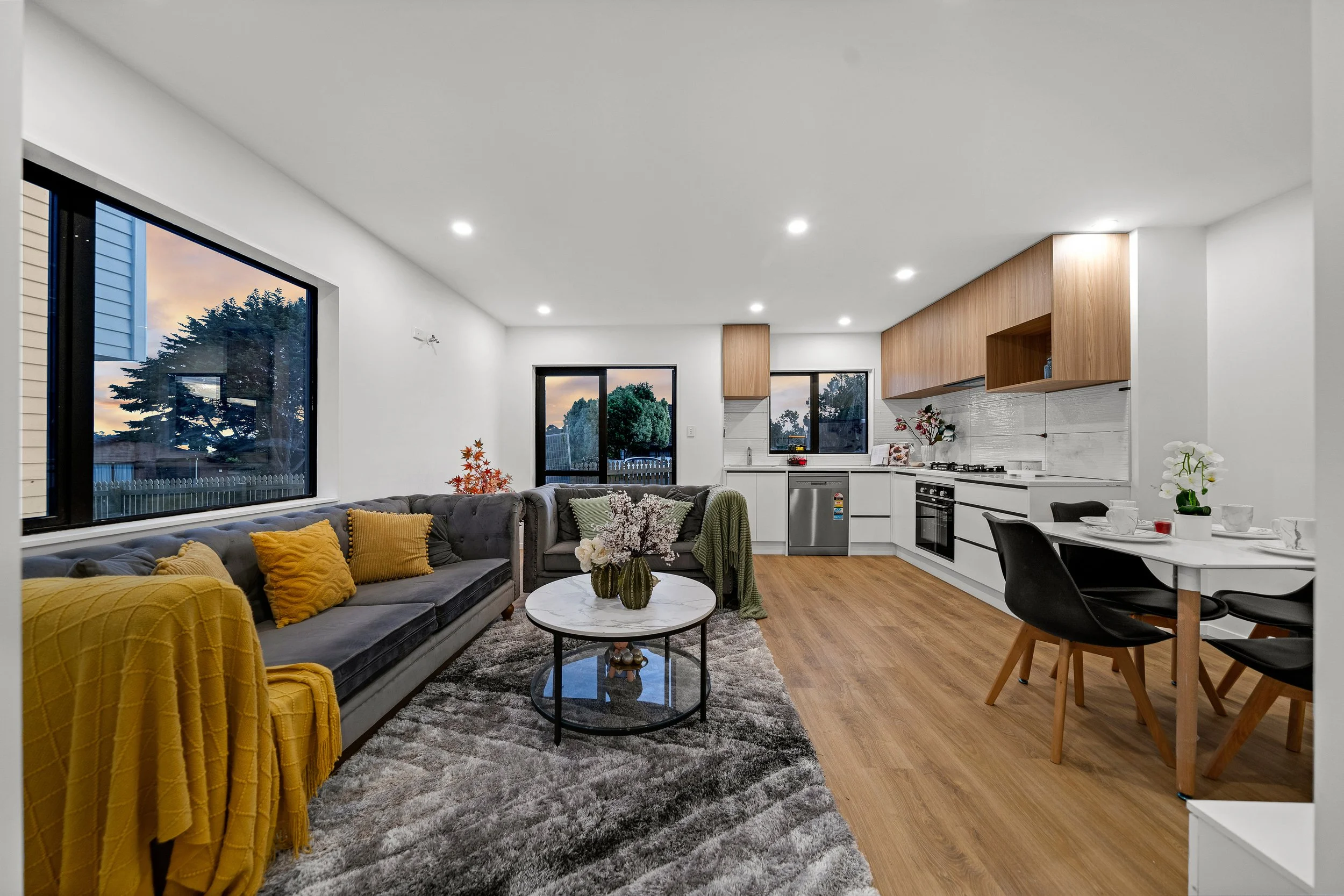 Modern open-concept living room and kitchen with large windows showing trees and a partly cloudy evening sky. The living room has a gray sofa with yellow and green pillows, a round marble coffee table with flowers, and a gray shag rug. The kitchen fe