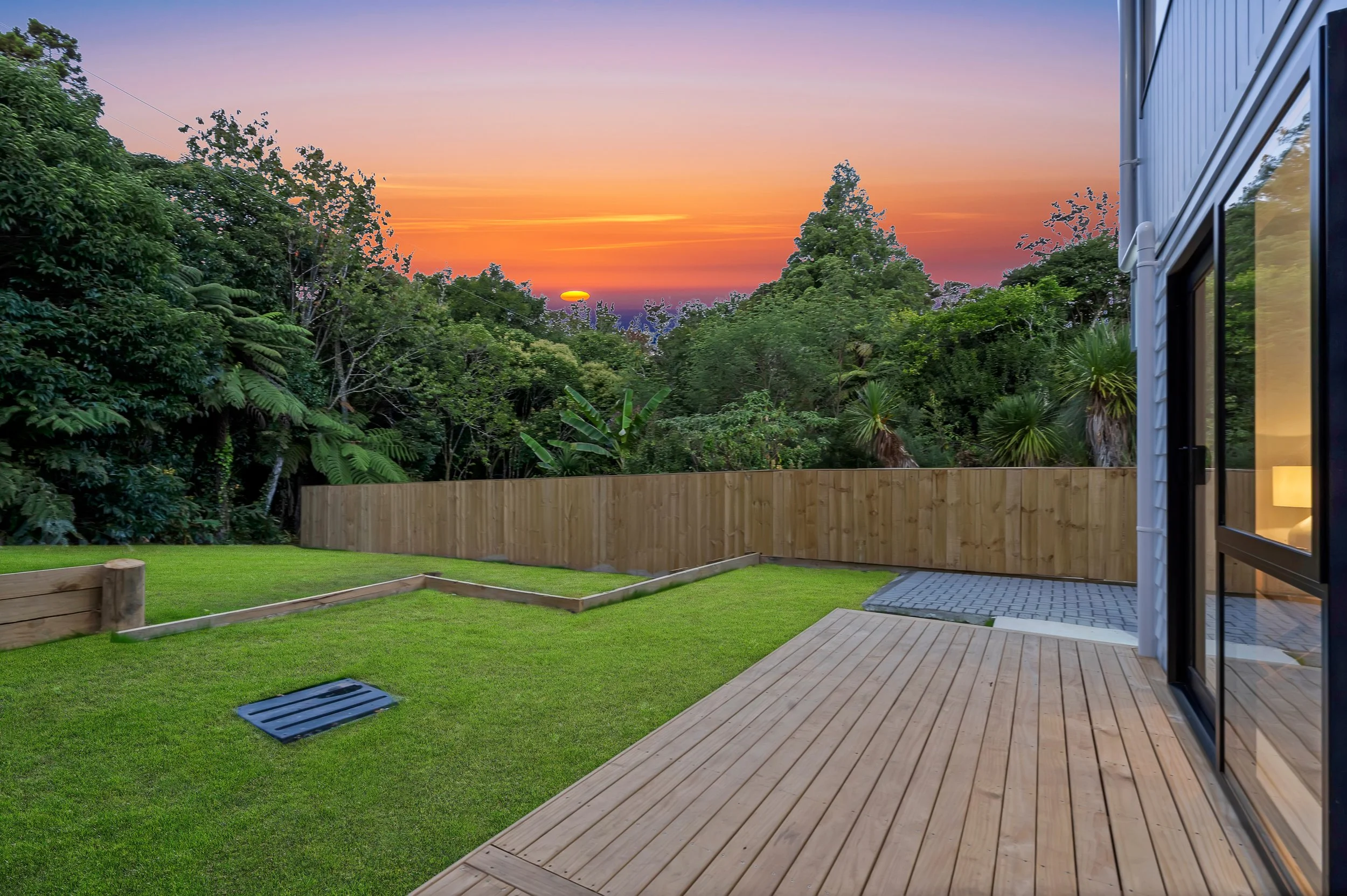 View of a backyard at sunset with a grassy lawn, wooden deck, and a wooden fence, surrounded by trees and foliage, with the sky showing orange, purple, and pink hues and the sun setting behind the trees.