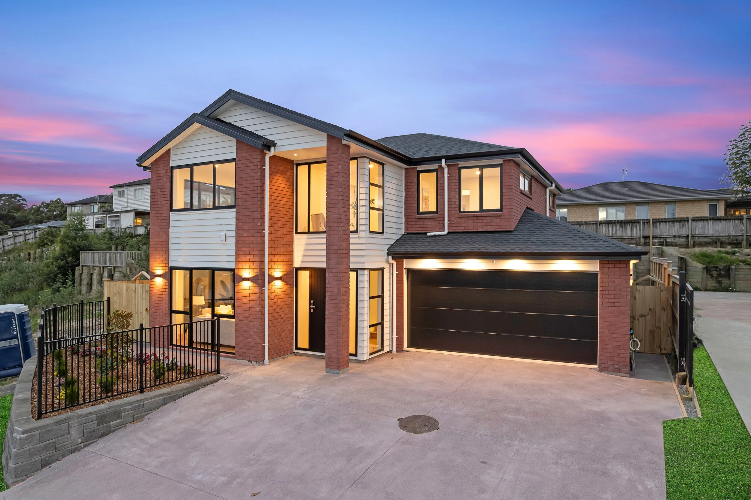 A modern two-story house with a brick and white siding exterior, large windows, and a black garage door, illuminated by outdoor lighting during sunset with colorful sky.