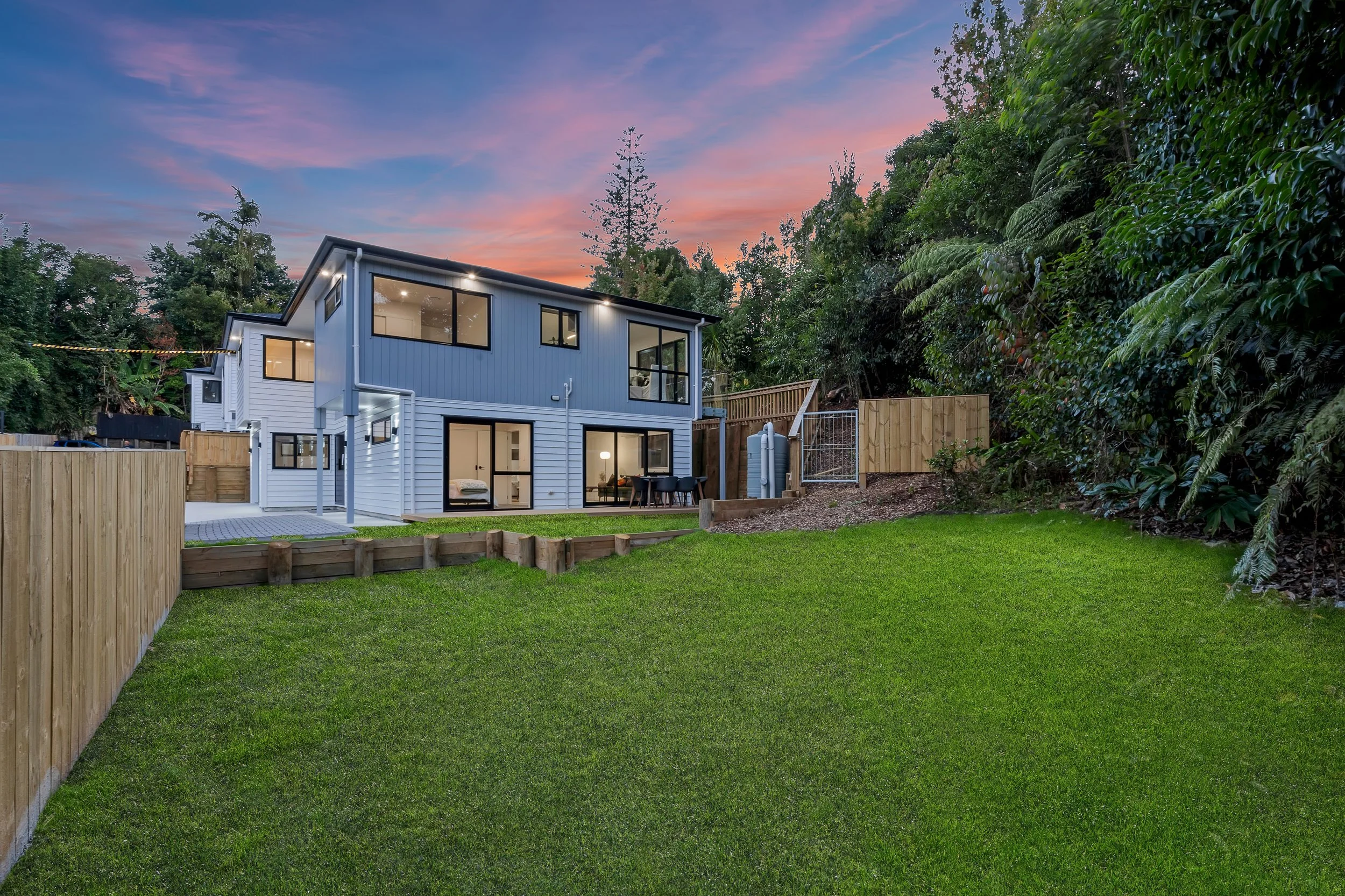 A modern two-story house with large windows, a backyard with a green lawn, a wooden fence, and surrounding trees during sunset.