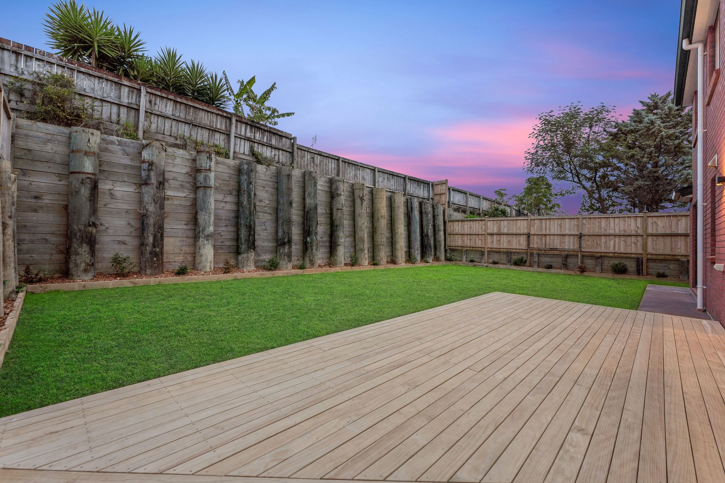A backyard with a wooden deck, green grass, wooden fencing, and trees; during sunset with pink and purple clouds.