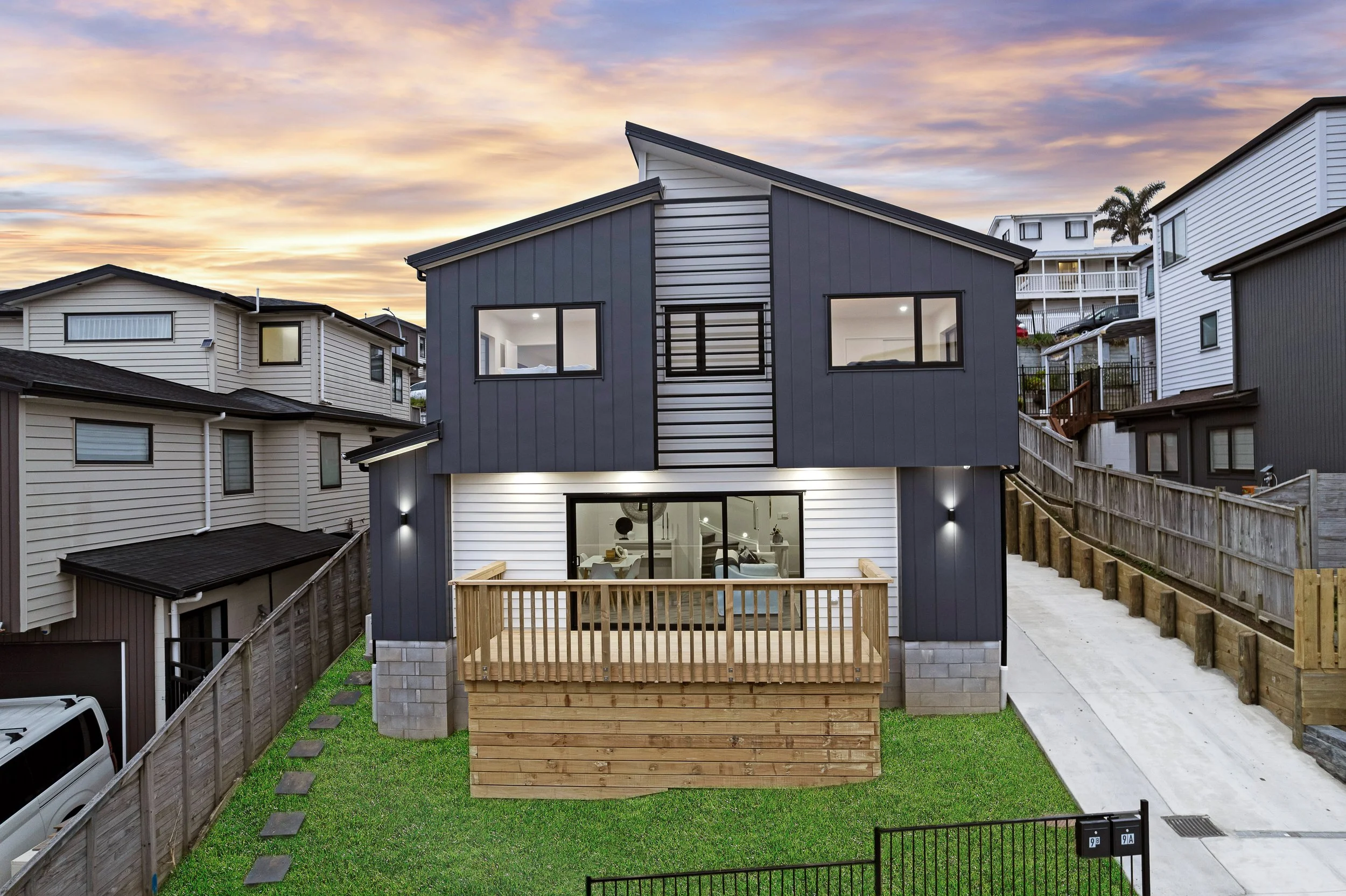 A modern gray and white two-story house with a small backyard deck, surrounded by fences and neighboring houses, during sunset with a colorful sky.