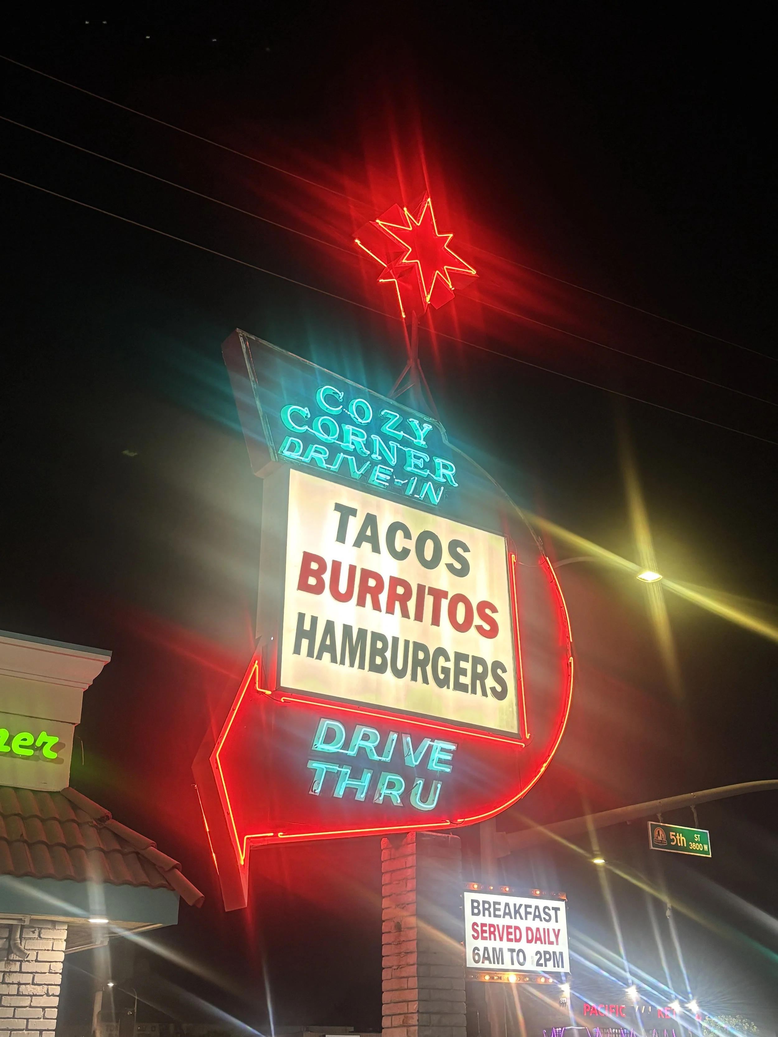 Neon sign advertising food items like tacos, burritos, and hamburgers, with a star and a smiley face, under a dark night sky.