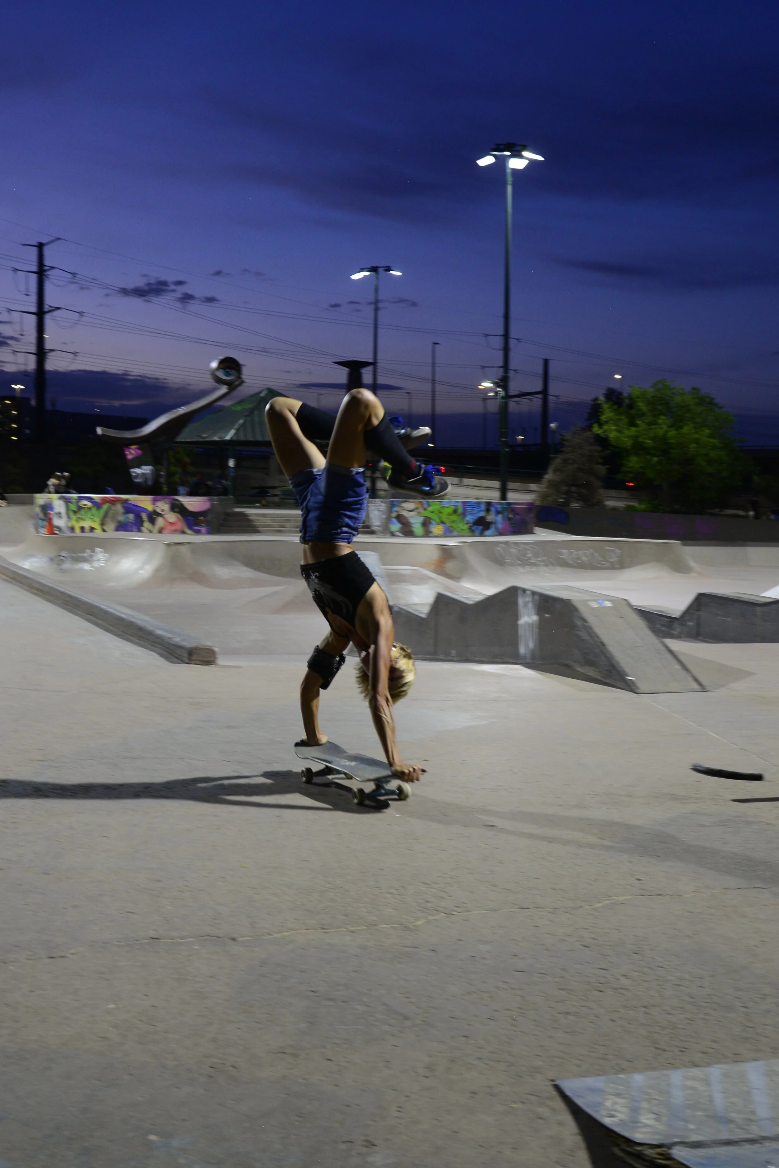 Person performing a handstand on a skateboard at a skatepark during twilight, with graffiti on the ramps and a dark cloudy sky in the background.