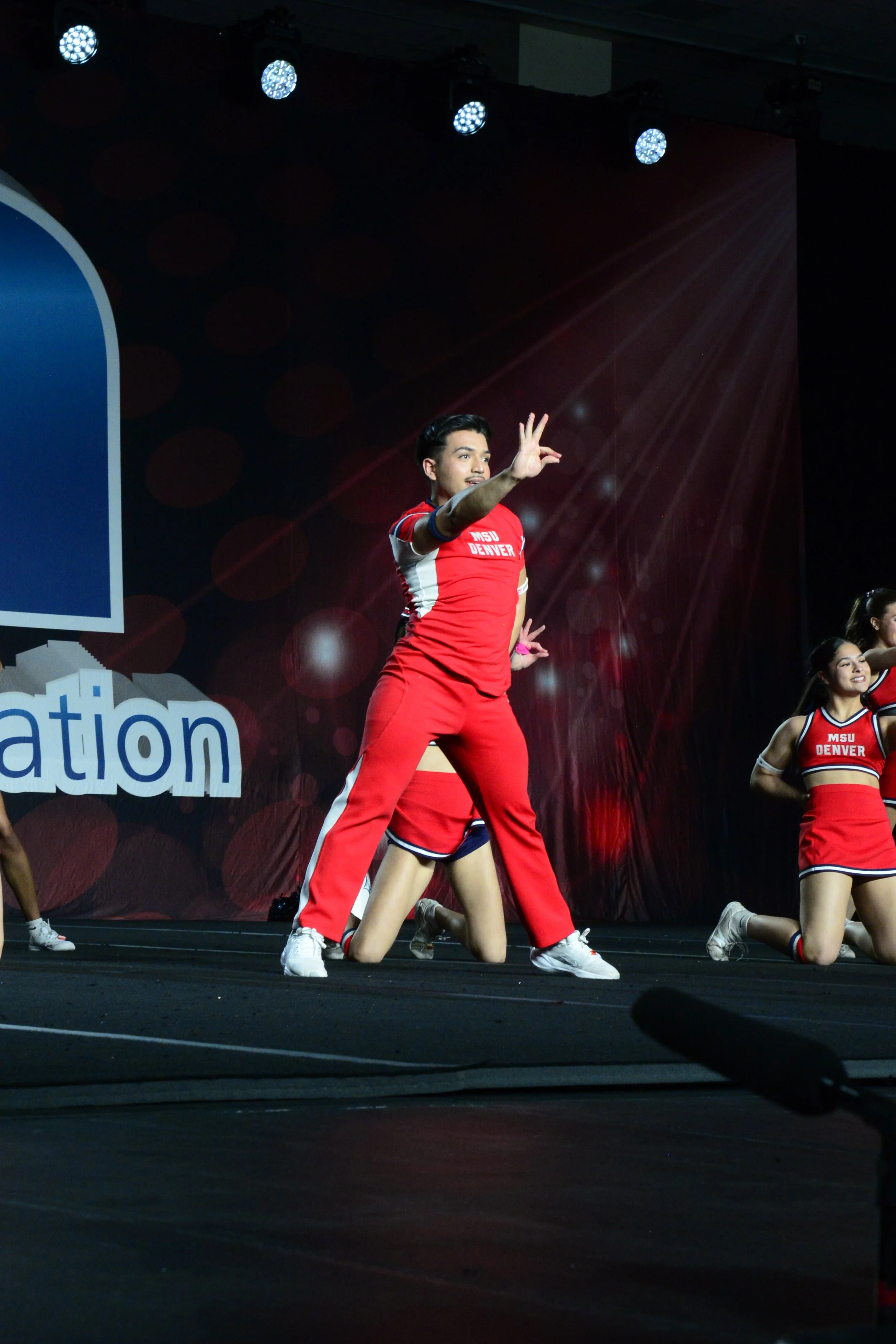 Cheerleaders performing a dance routine on stage, wearing red and white uniforms that say "MSU Denver" and "Cheer".