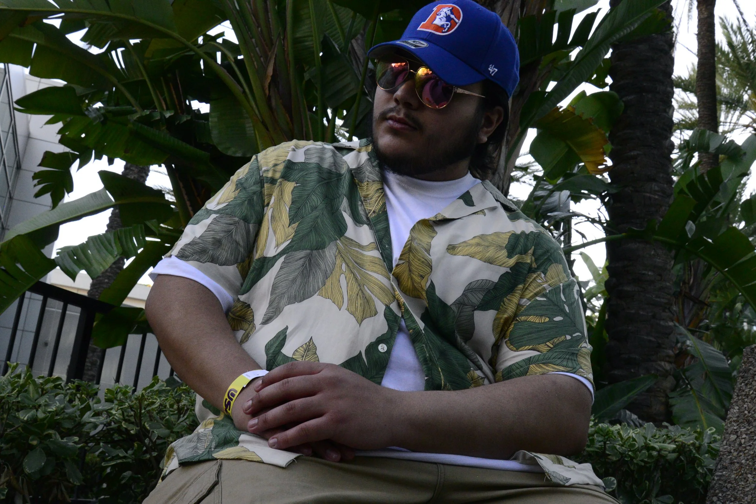 A man wearing sunglasses, a blue cap with a logo, a tropical print shirt, and beige pants, standing outdoors among large green tropical plants and palm trees.
