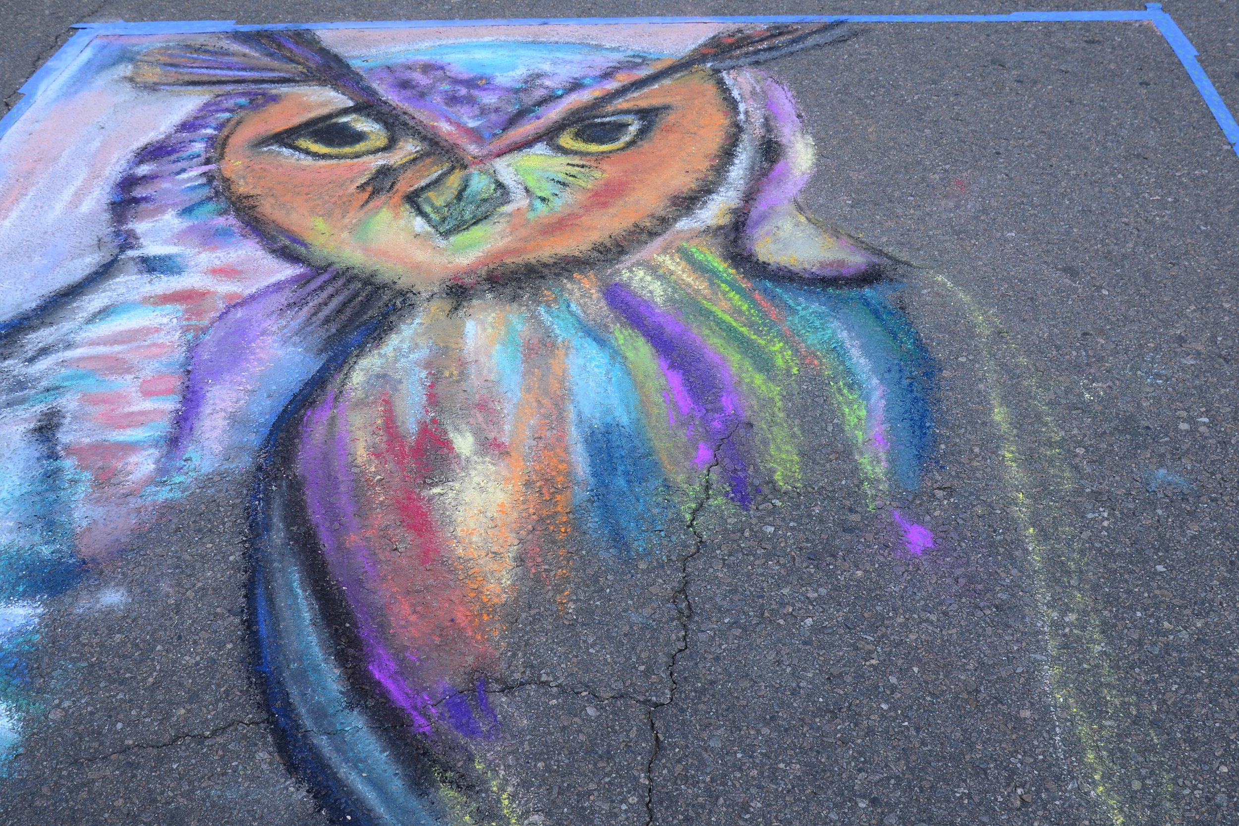 Colorful chalk drawing on pavement depicting a stylized, expressive face with intense eyes and vibrant rainbow colors.