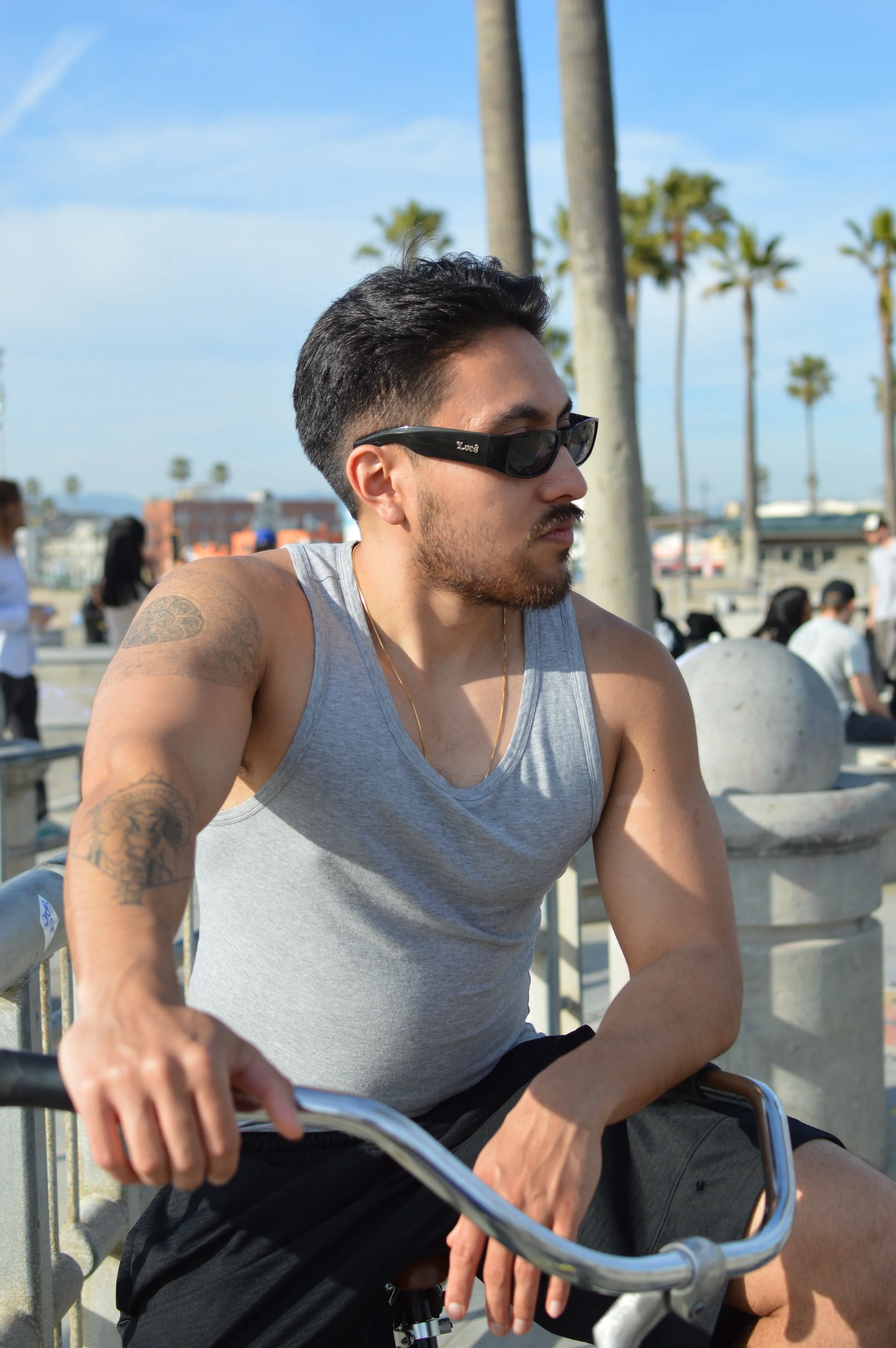 A man with short dark hair, sunglasses, a beard, and tattoos on his arm, wearing a gray tank top, sitting outdoors on a sunny day with palm trees in the background.