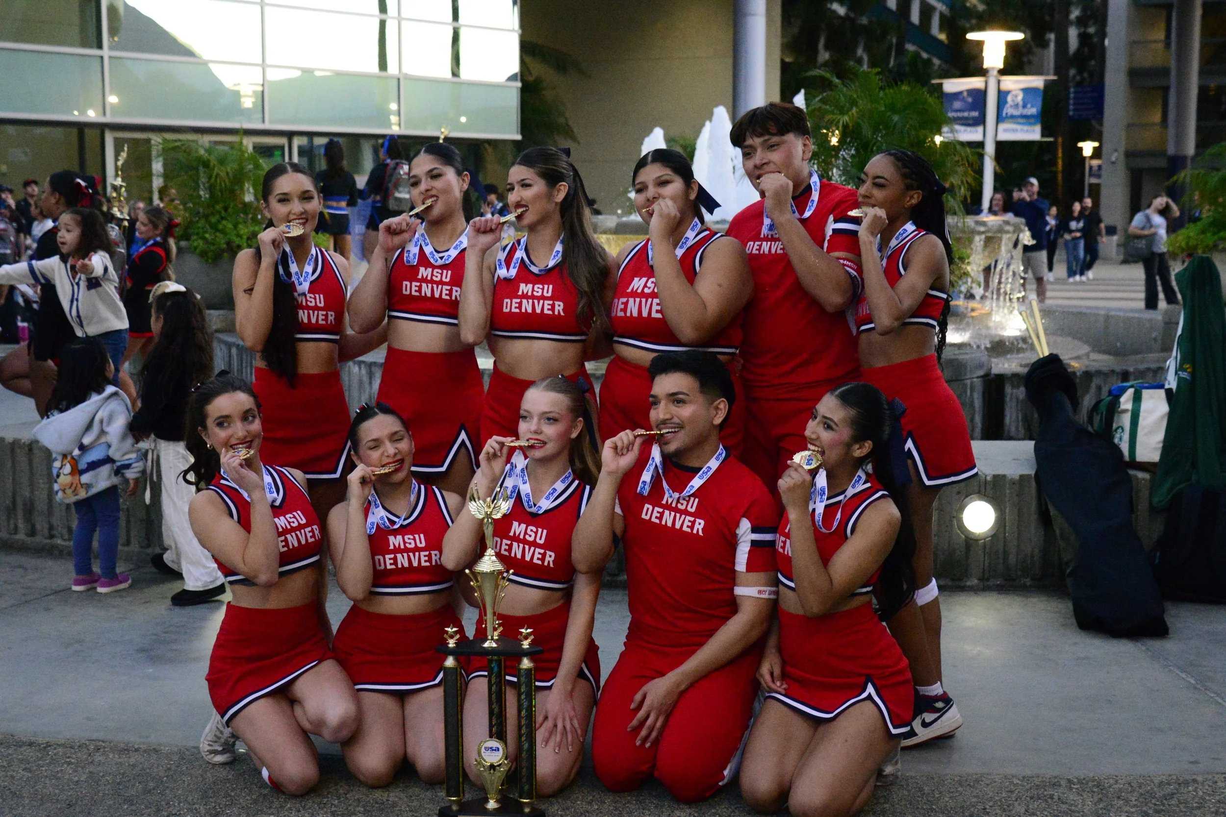 A group of cheerleaders in red and white uniforms holding medals and a trophy, celebrating their victory outside in front of a fountain.