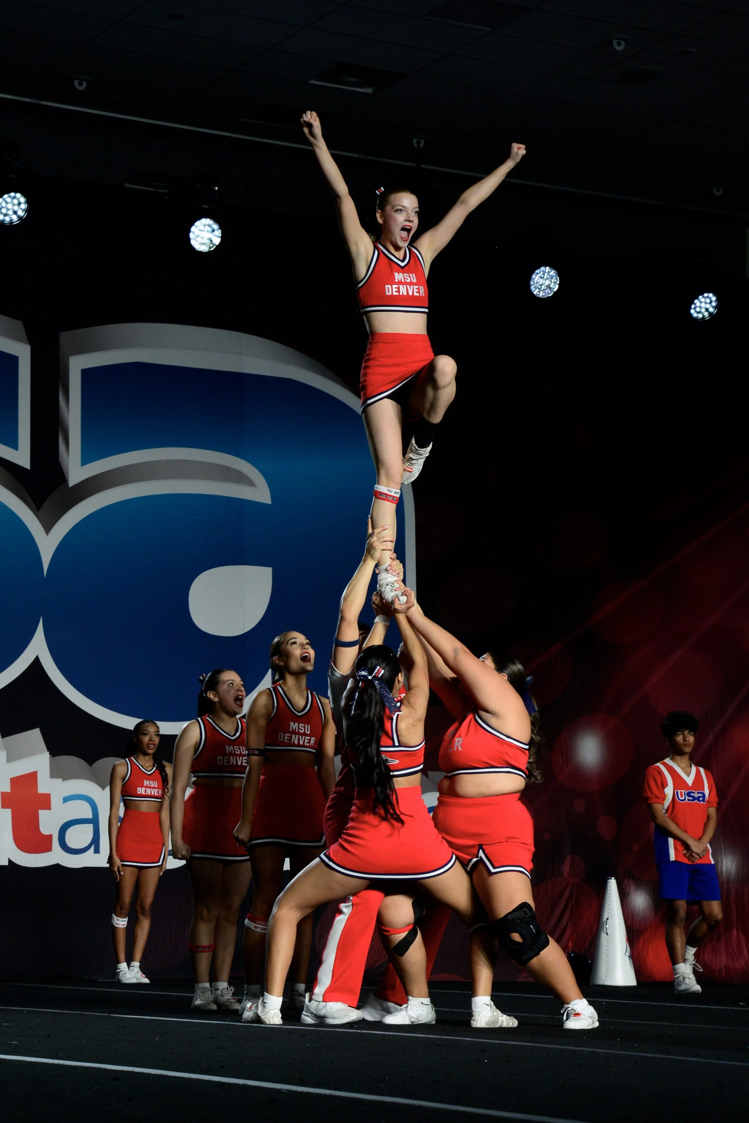Cheerleaders in red uniforms forming a pyramid during a performance at a gym or stage, with a cheerleader at the top raising her arms and others at the base holding her up.