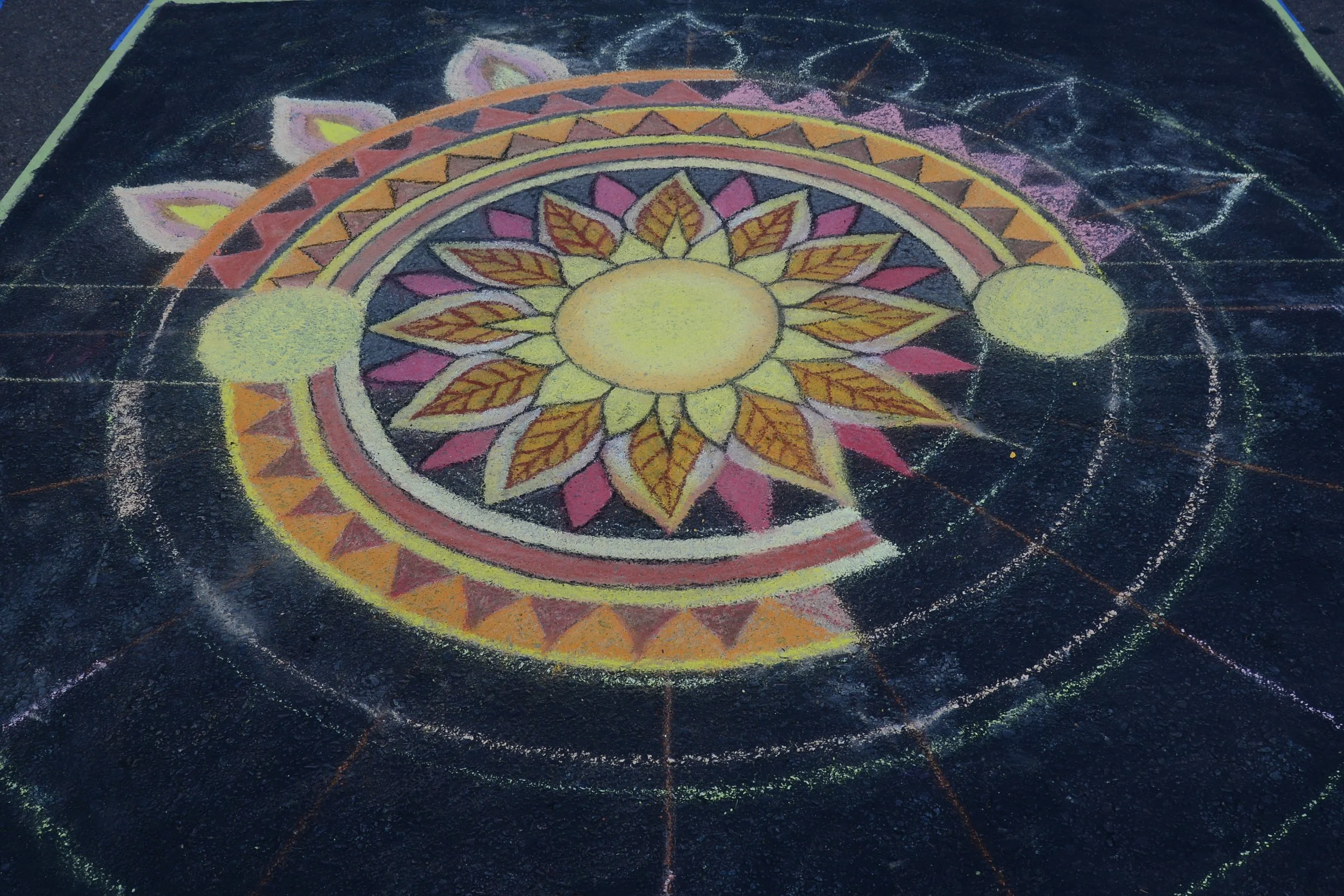 Colorful chalk drawing of a mandala on the ground, featuring a central yellow circle surrounded by leaves, geometric shapes, and floral patterns with vibrant pink, orange, yellow, and purple colors, on a black asphalt surface.