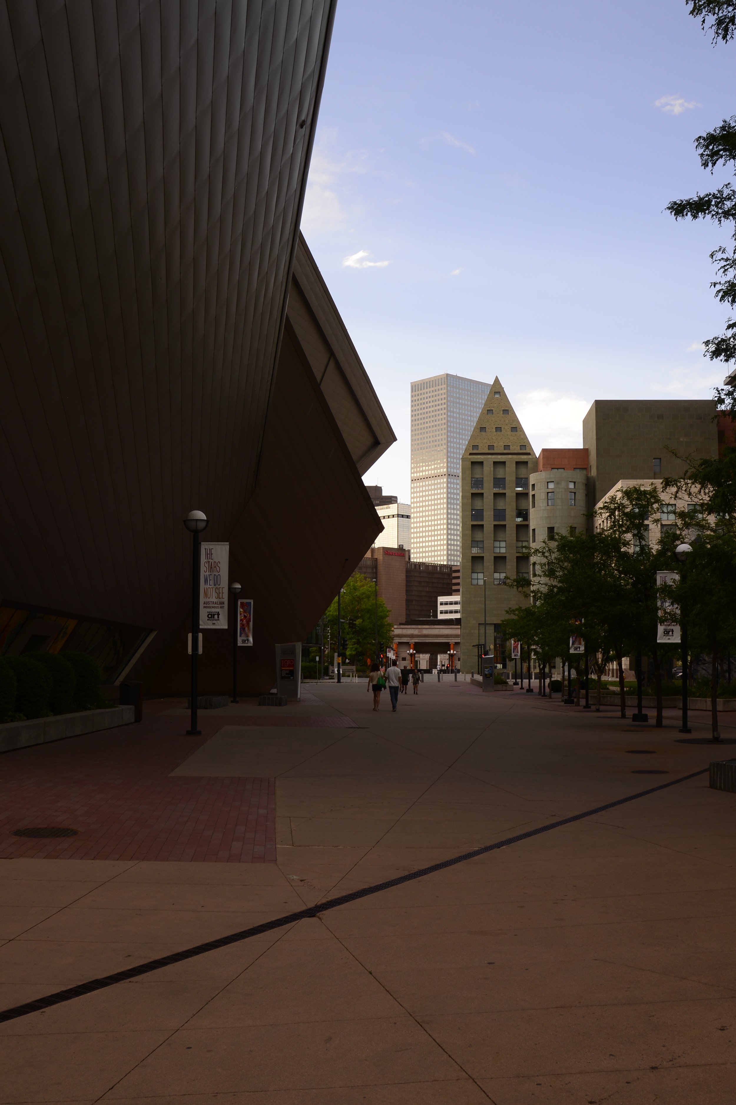 A city street scene with tall buildings in the distance, some trees on the right, and a large modern architectural structure with a sloped roof on the left. There are a few pedestrians walking down the sidewalk.