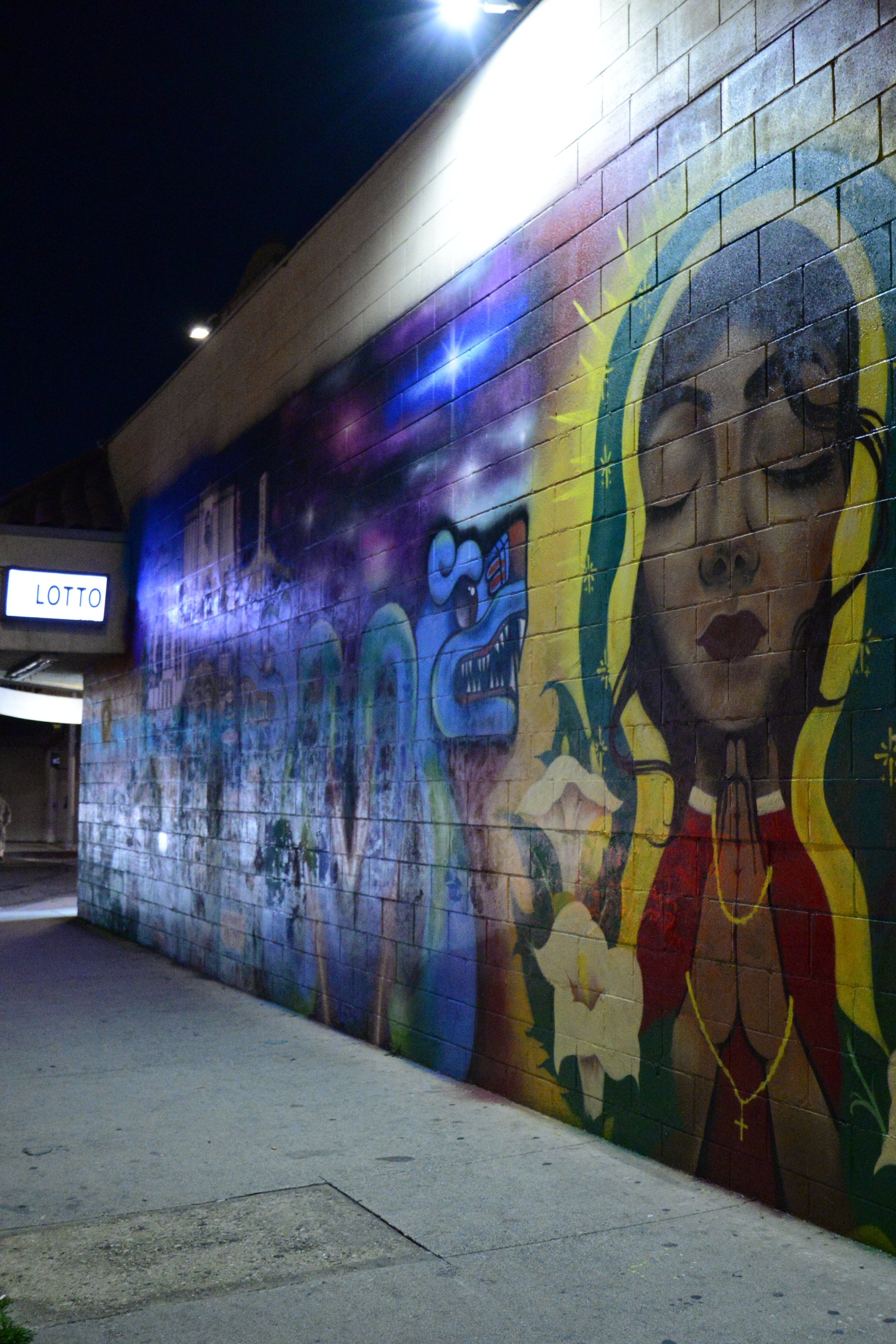 Nighttime mural on a brick wall depicting a stylized portrait of a woman with long, flowing hair and closed eyes, illuminated by a streetlight.