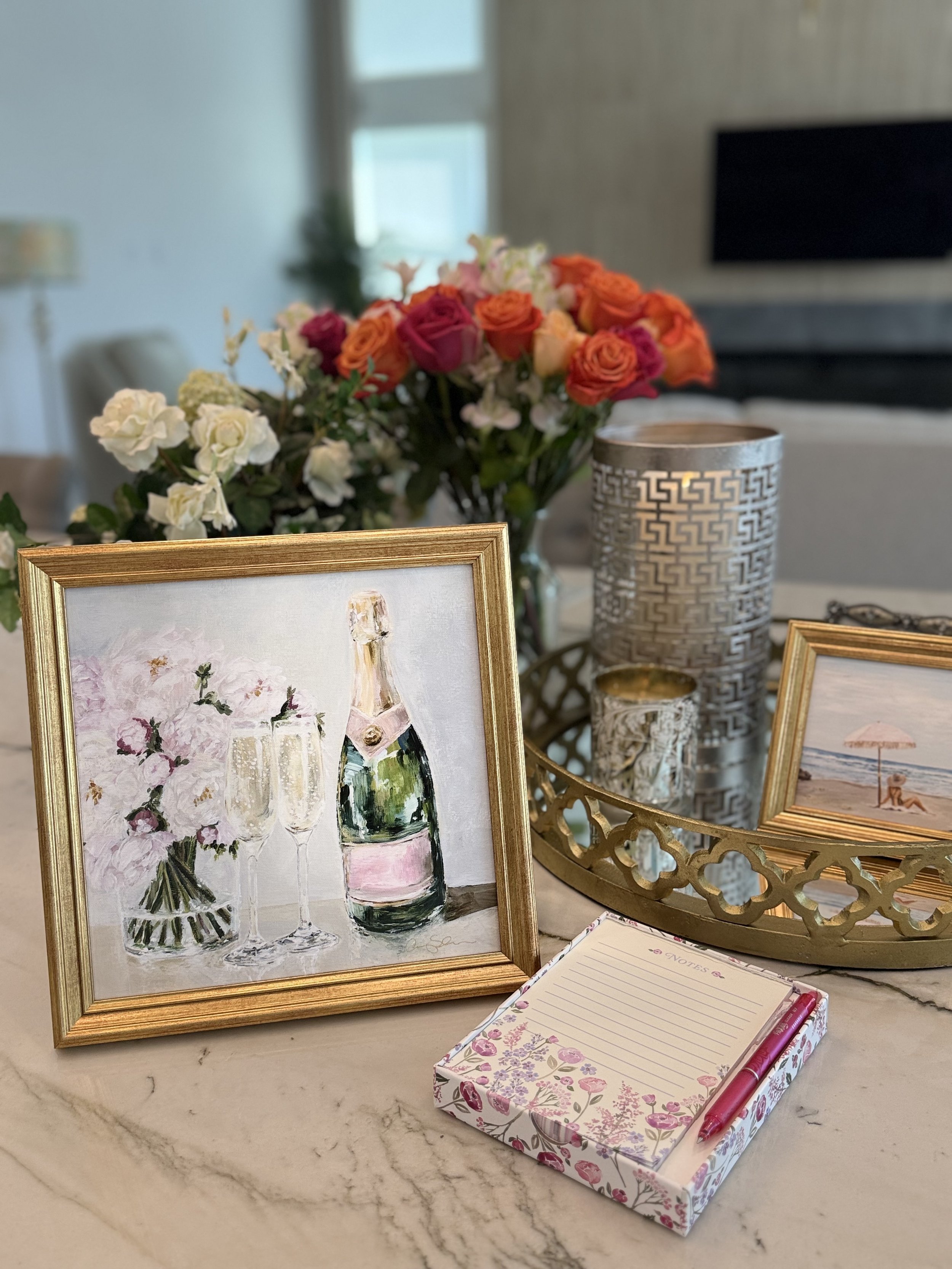 A decorated table with framed watercolor paintings of champagne glasses and flowers, a silver patterned candle holder, a small candle, and a pink floral notepad with a pink pen.