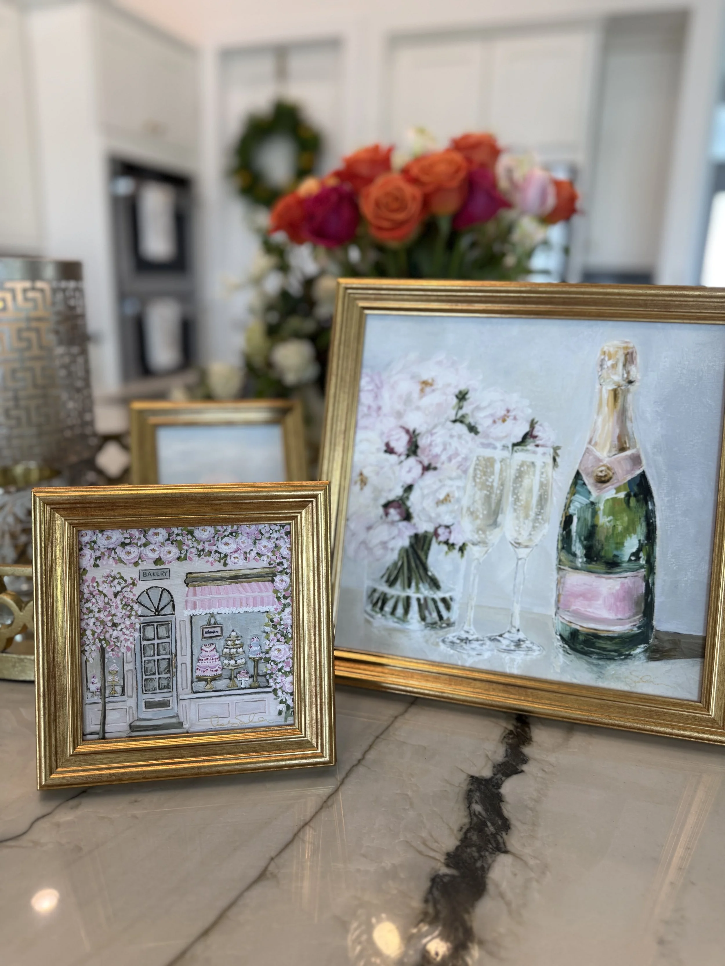 Two framed paintings of floral scenes on a marble surface, with a bouquet of pink and orange roses and white flowers in the background, inside a room with a white wall and a decorative wreath.