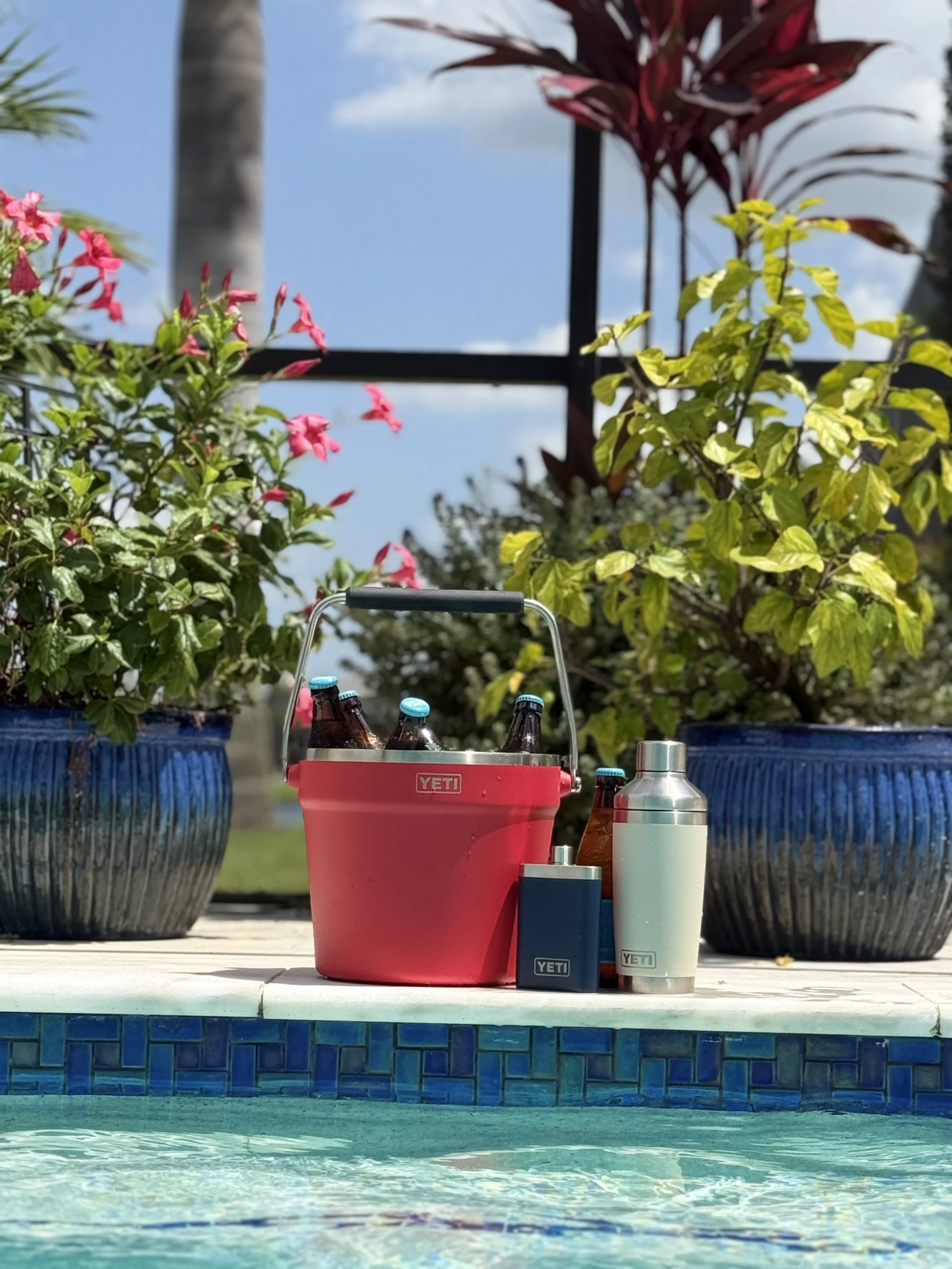 A poolside scene with two large blue pots with plants, a red YETI cooler with six bottles inside, and various other bottles and containers on a white surface, with a background of clear sky and palm trees.