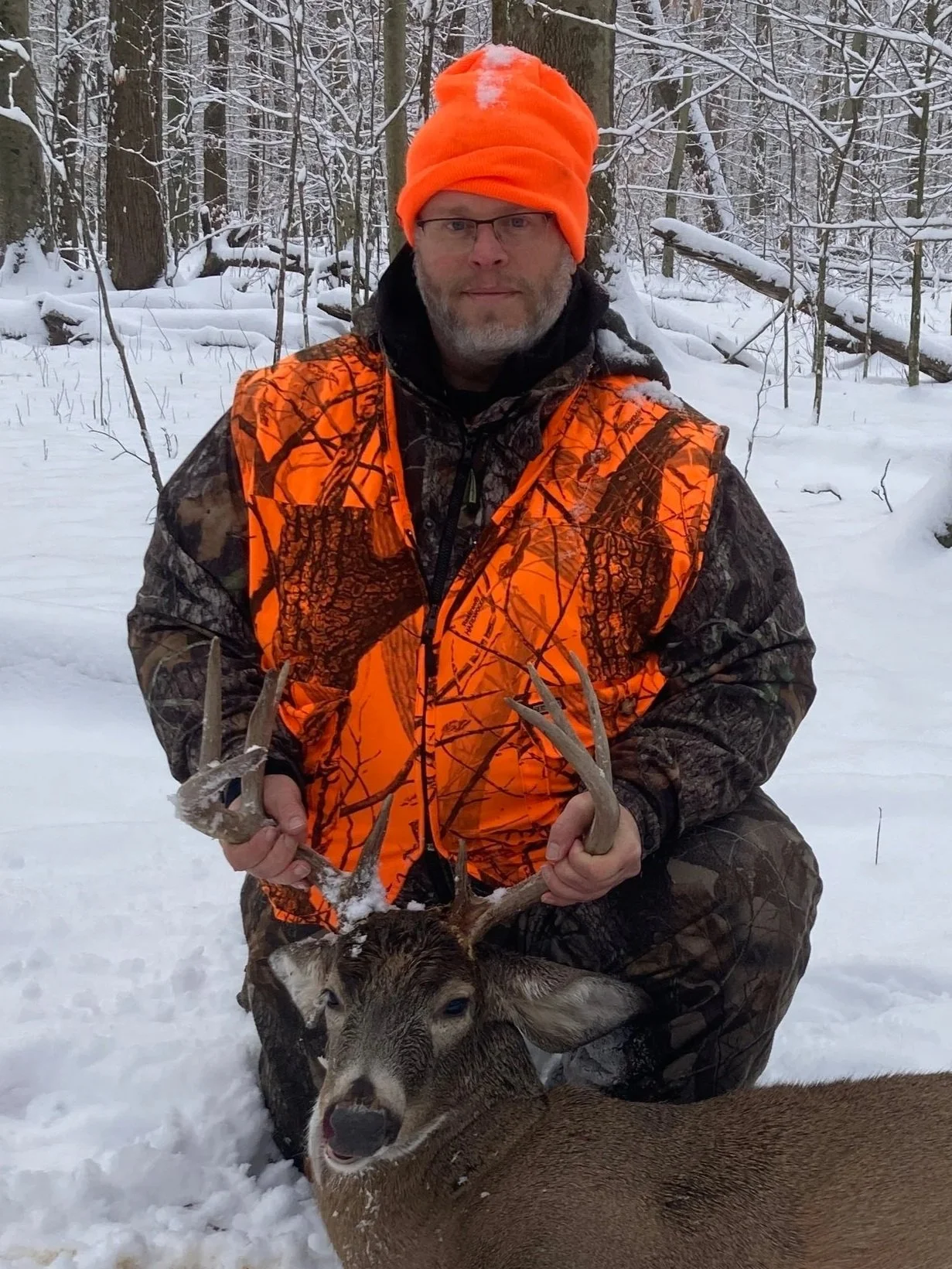 A man in camouflage and bright orange hunting gear kneels in a snowy forest, holding a deer with antlers. The man wears glasses and an orange knit hat, and the deer is lying on the snow.