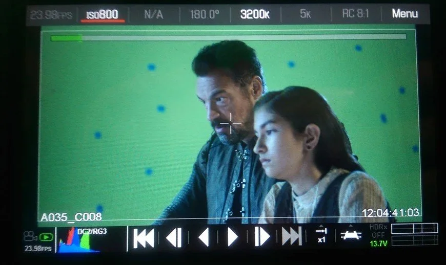 Two people, a man and a woman, are sitting in front of a green screen with blue dots. The man is looking slightly to the left, and the woman is looking straight ahead. They appear to be on a film or video production set.