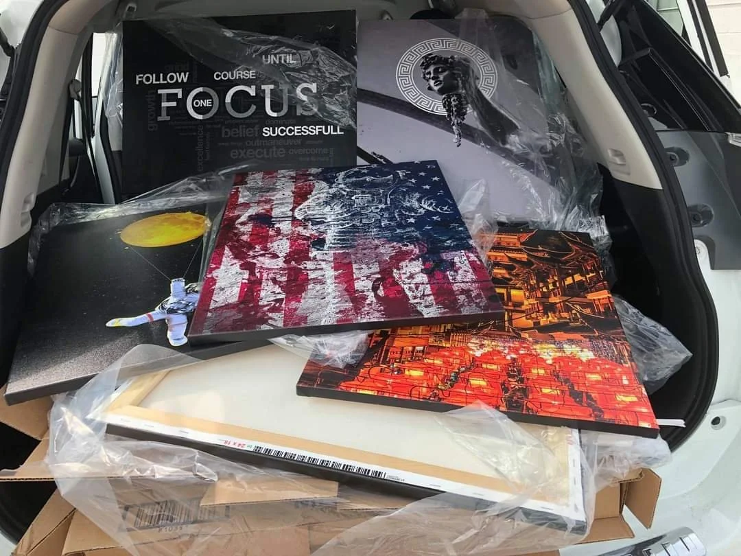 The trunk of a car filled with framed wall art and posters, including space-themed artwork, a black and white poster with the words "Follow your course until you focus. Successful." and an image of Medusa.