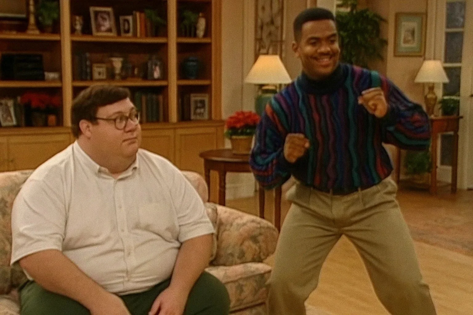 A man with glasses and a white shirt sitting on a floral patterned couch while another man in a colorful striped shirt and khaki pants stands in a lively pose next to him in a living room with wooden bookshelves, lamps, and framed pictures on the wal
