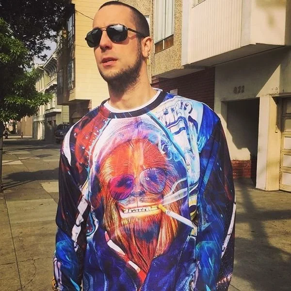 A man wearing sunglasses stands on a city sidewalk, dressed in a colorful long-sleeve shirt with a graphic of a dog with sunglasses and a cigarette.