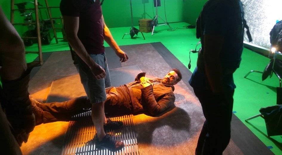 Man lying on a green screen studio floor surrounded by crew members, with lighting equipment in the background.