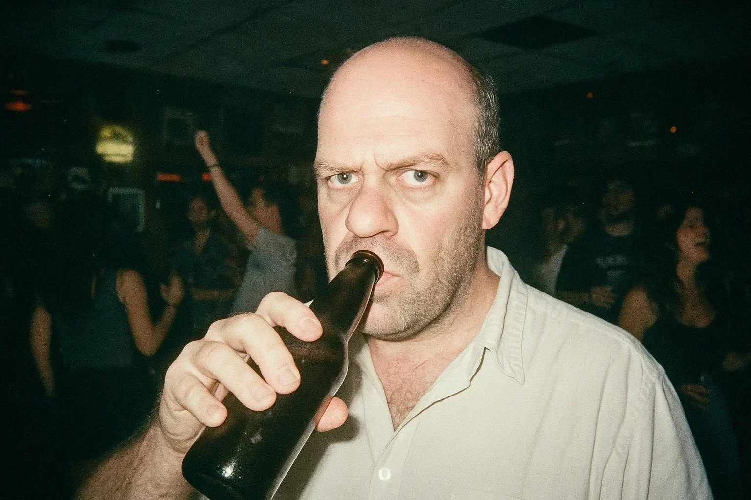 A man with a serious expression drinking from a dark bottle at a crowded indoor venue.