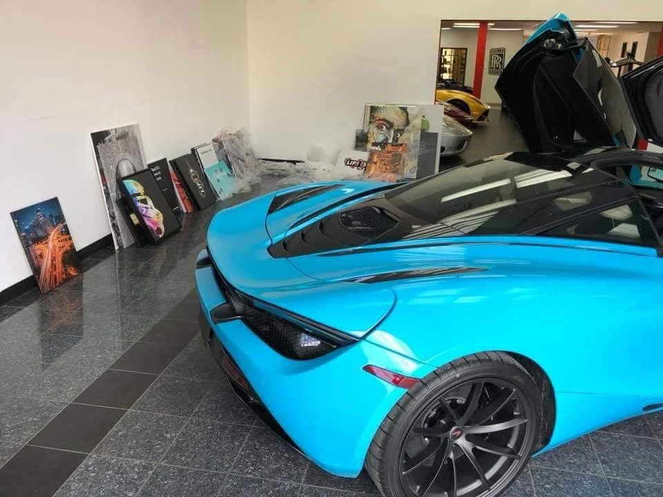 Blue sports car with its gullwing door open in an indoor showroom, next to a row of various colorful paintings and artwork leaning against the wall.